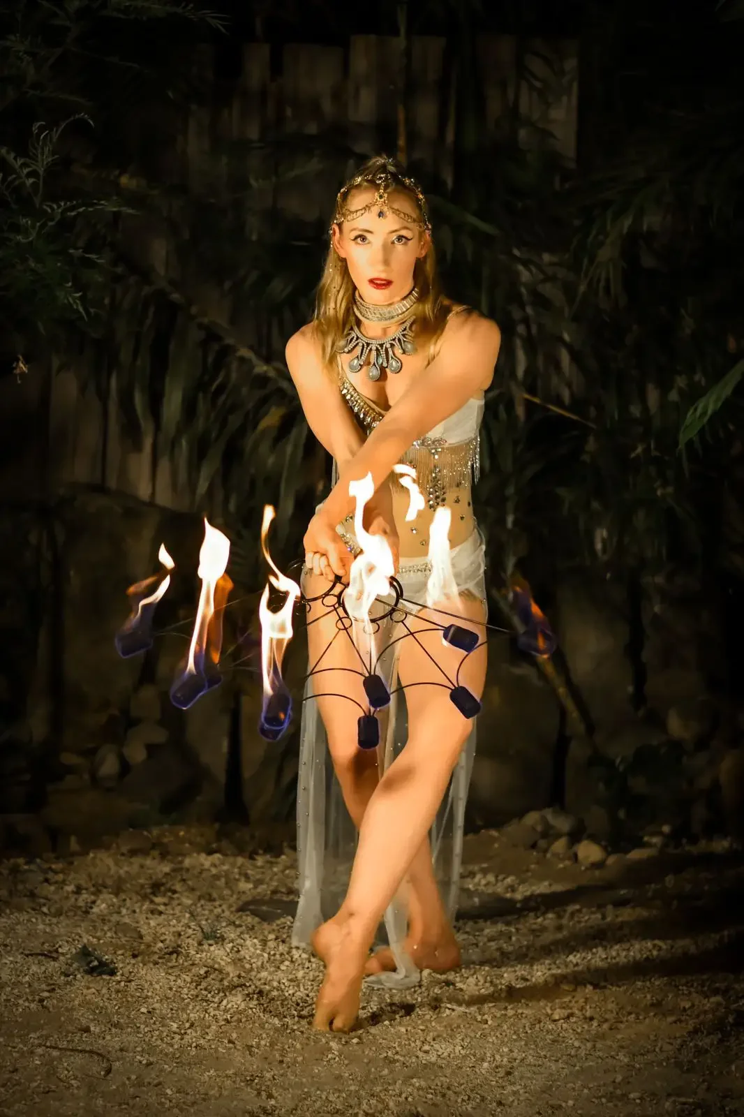 A woman performing a fire dance outdoors at night, holding fire poi in front of a dark background with greenery.