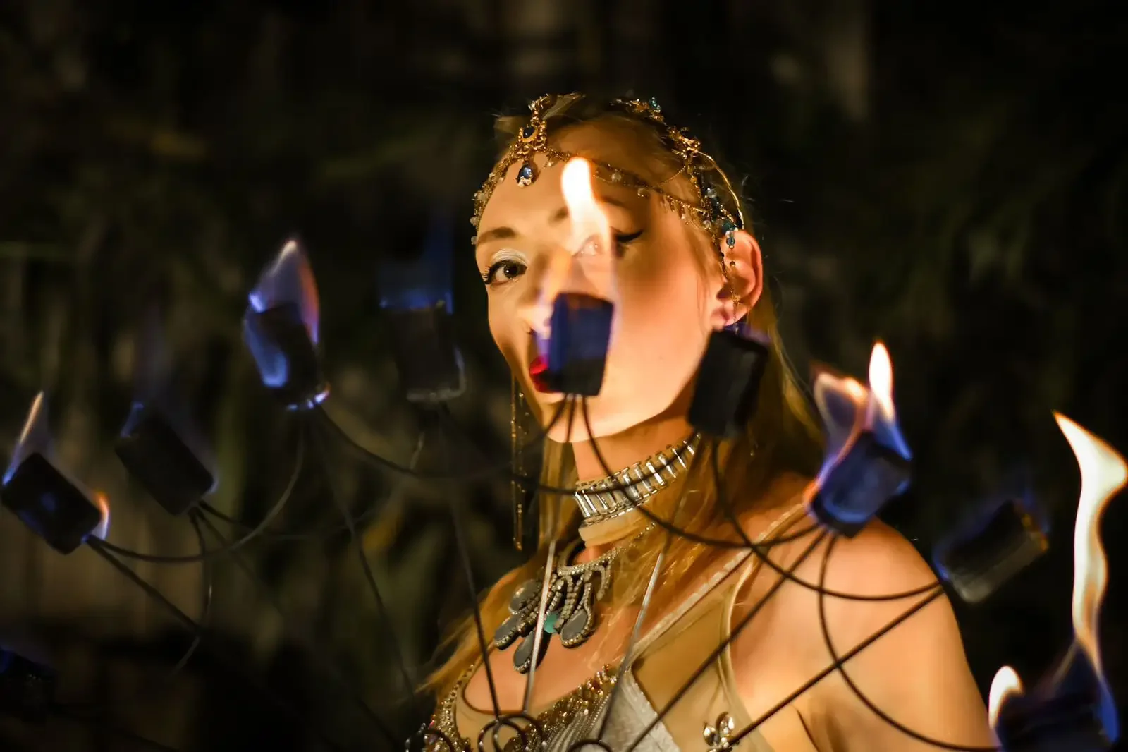 A woman wearing jewelry and makeup, with a headpiece, stands amid flames, holding a fire poi leading a fire performance.