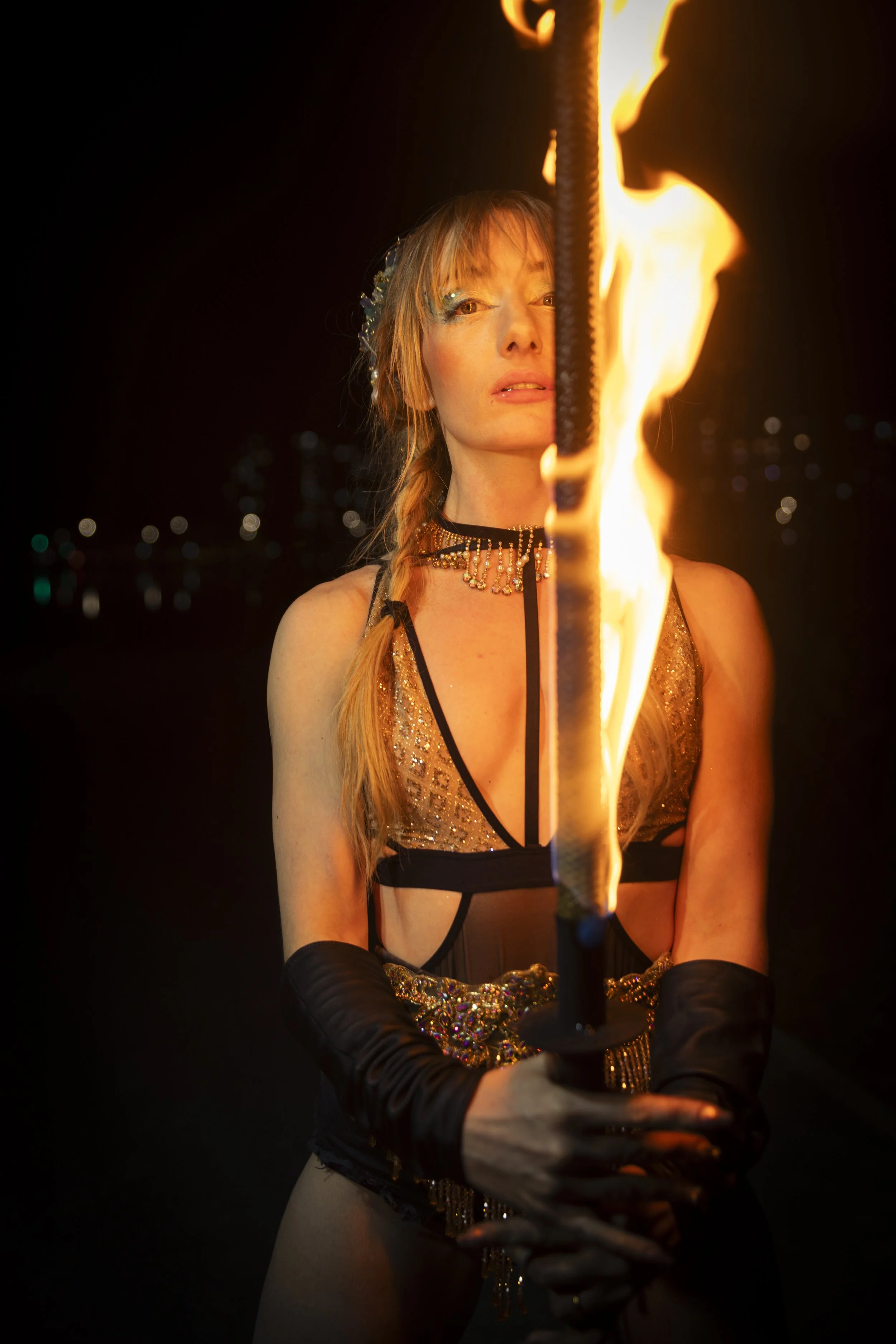 A woman with long blond hair in a braid holding a flaming torch at night, dressed in a sparkly gold and black outfit with long black gloves.