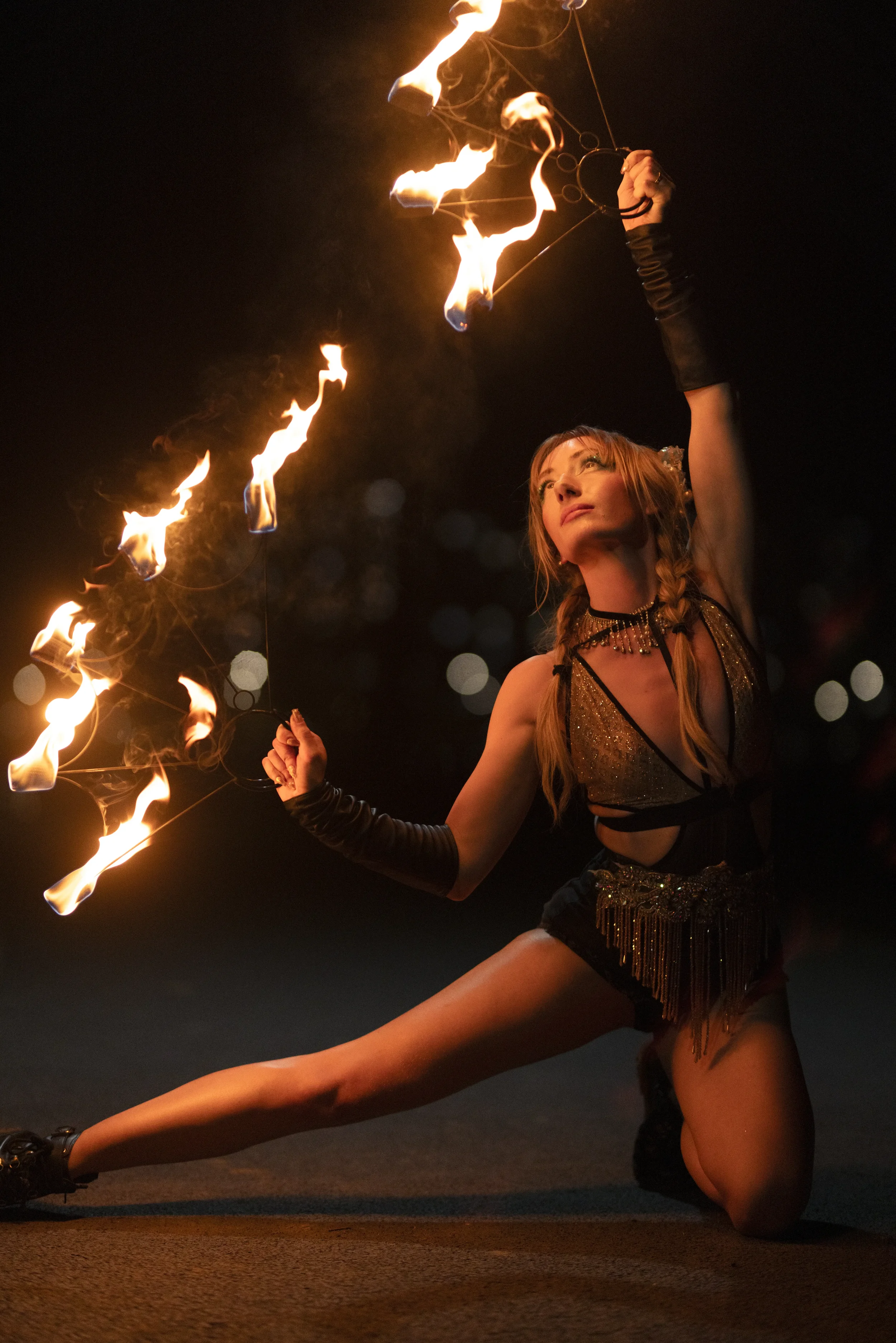 A female fire dancer performing at night, holding fire poi in both hands with flames in motion, wearing a sparkly outfit and long gloves.