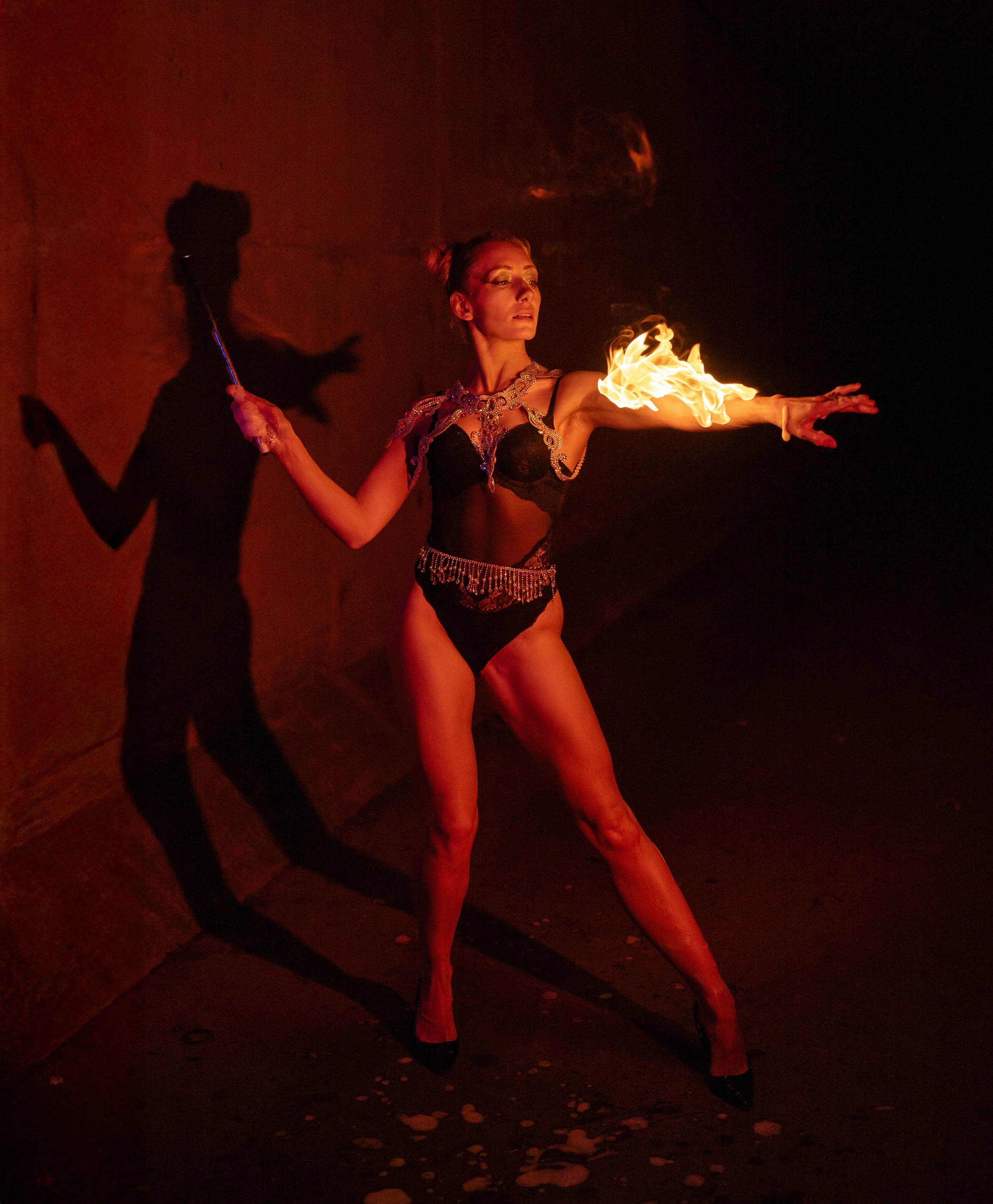 A female fire performer in a black outfit with gold embellishments holds a flaming torch, with her arm extended and fire on her hand, casting a shadow on the wall behind her, in a dark environment.