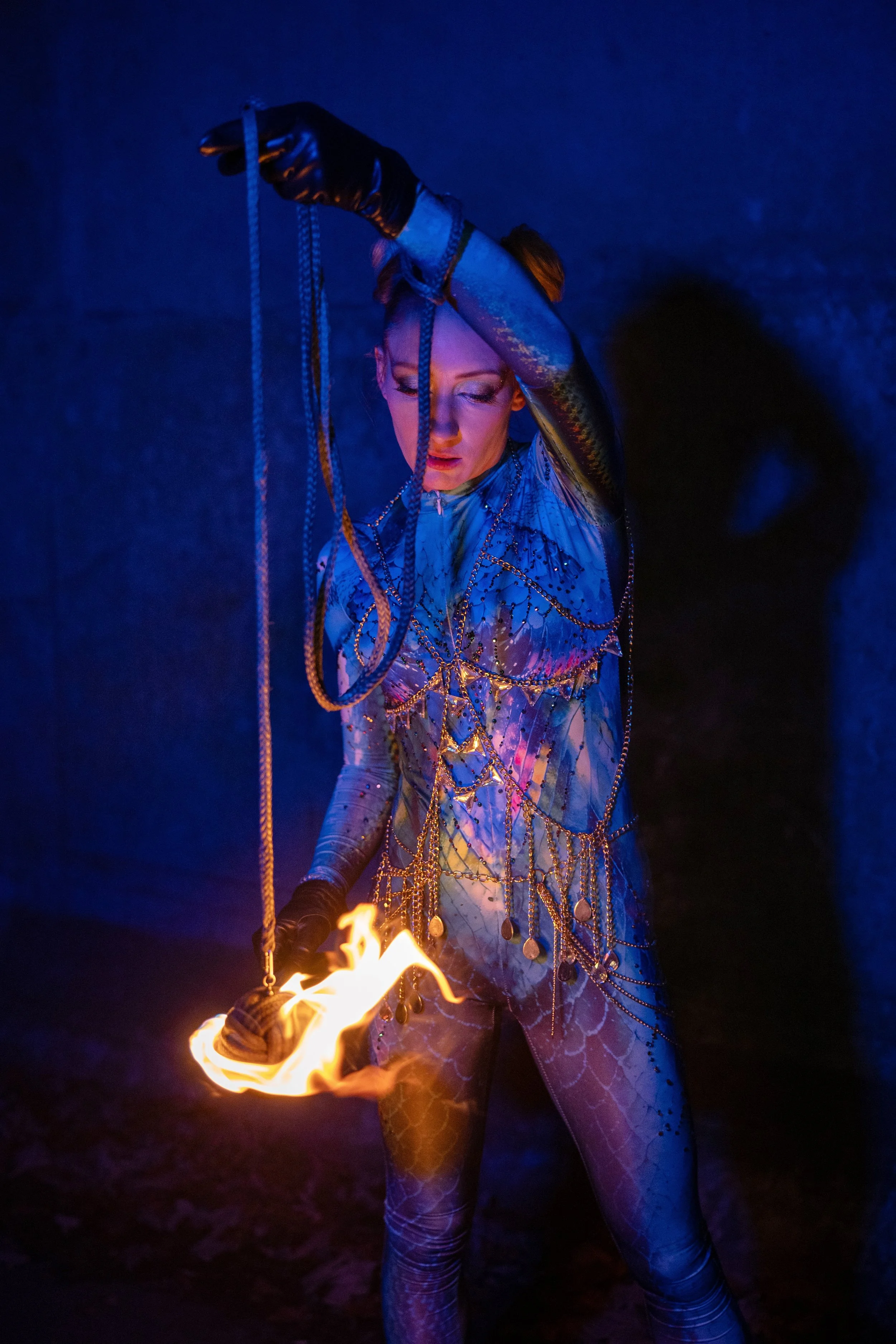 A woman wearing a bodysuit with a blue and purple pattern and gold chains draped over her, holding a flaming object in one hand, with a dark background and a cloud of smoke in the air.