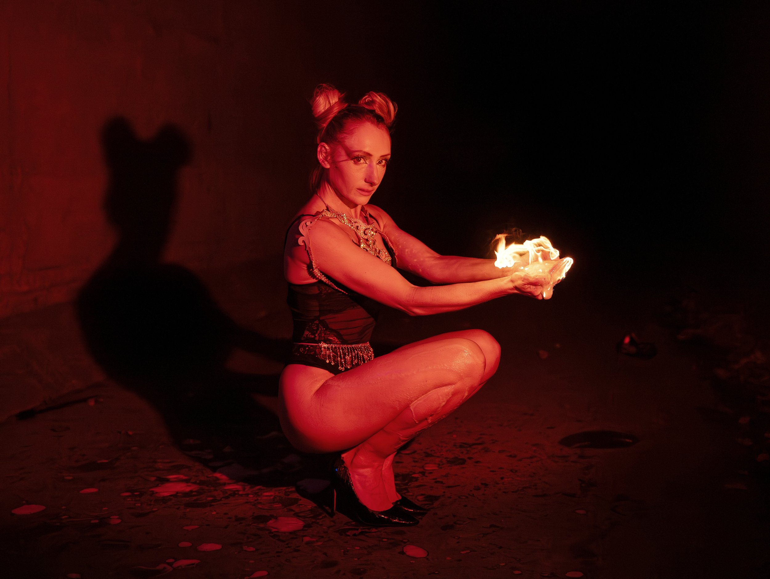 A woman with blonde hair styled in two buns, squatting in a dimly lit space, holding a fireball in her hands, with her shadow cast on a brick wall behind her.