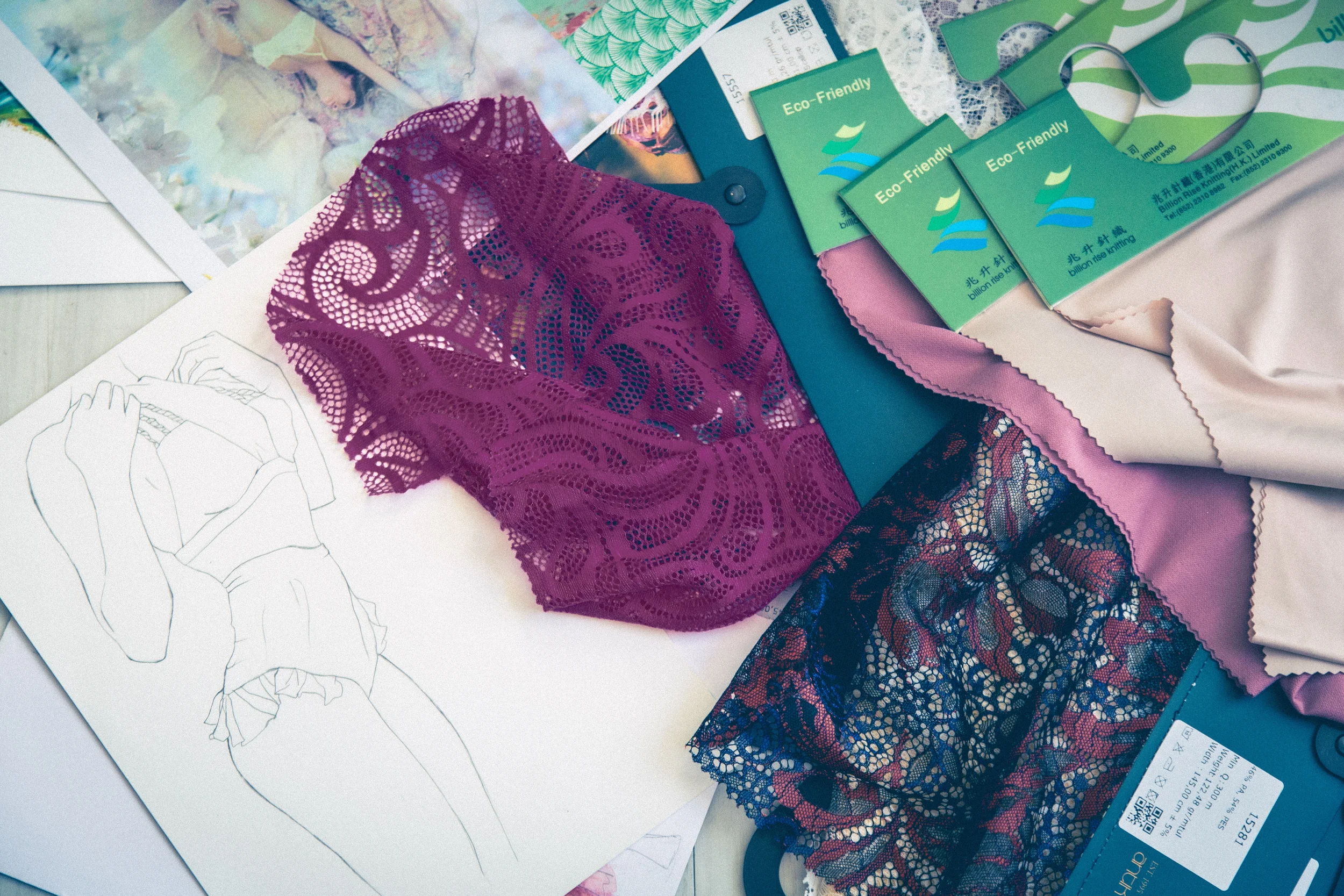 A flat lay of lingerie fabric swatches, lace trim, and a fashion illustration sketch on paper. An illustration of a swimwear design in pencil also features.