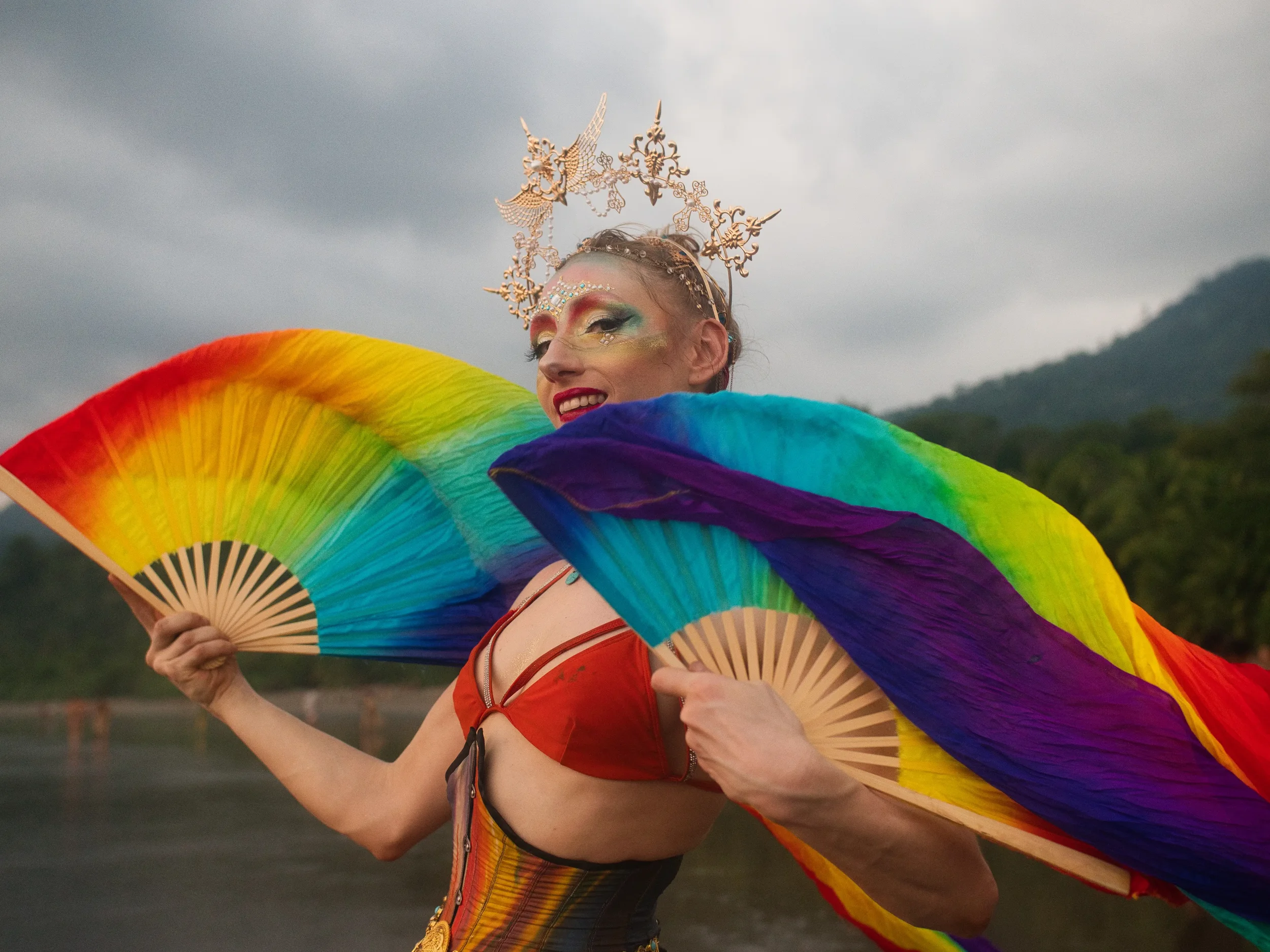A person dressed in colorful pride attire holding rainbow-colored fans, wearing an elaborate headpiece with gold designs, makeup, and smiling outdoors with cloudy sky and mountains in the background.