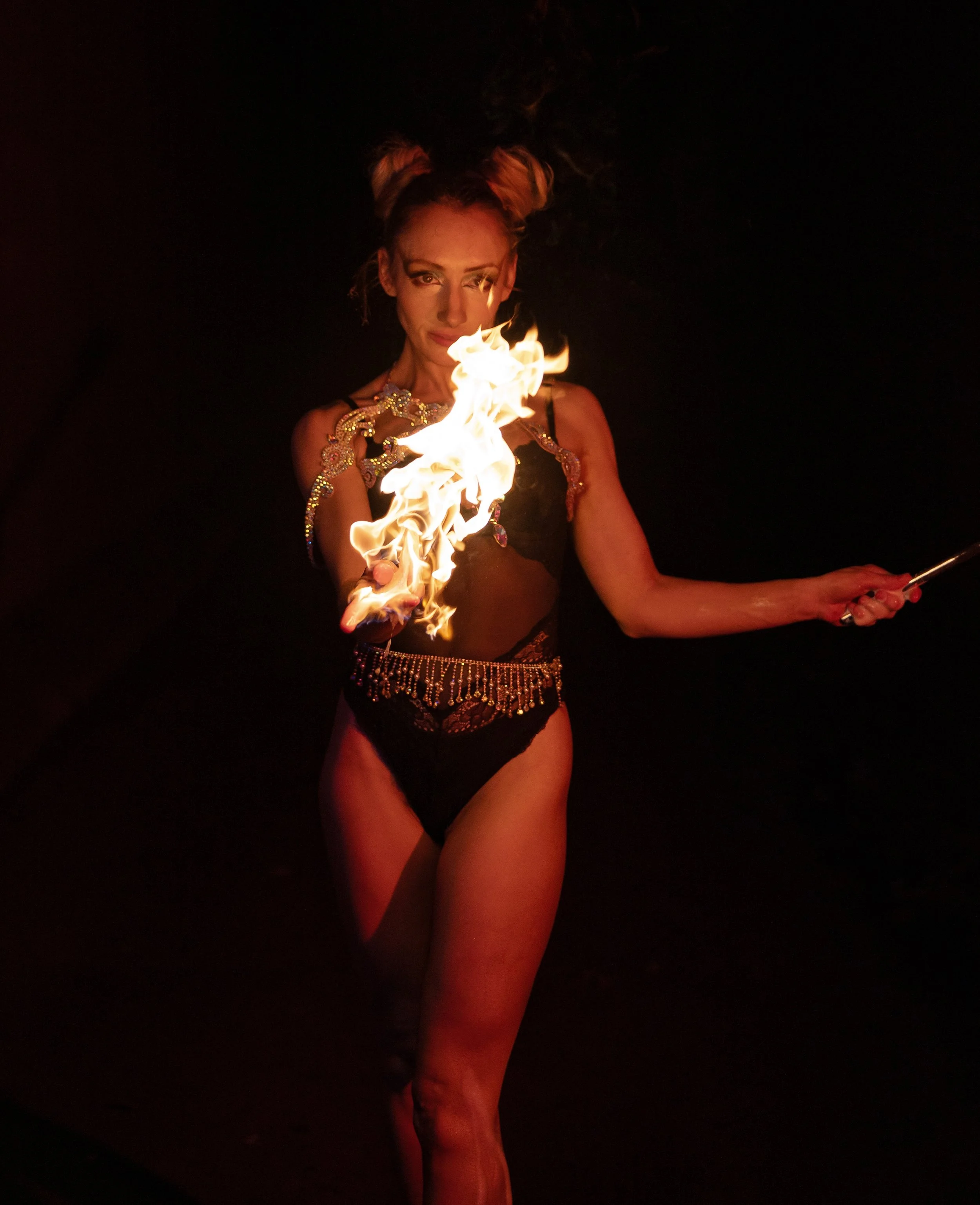 A woman in black lace clothing performs a fire-eating act, holding a flaming torch in one hand with fire near her face in a dark setting.