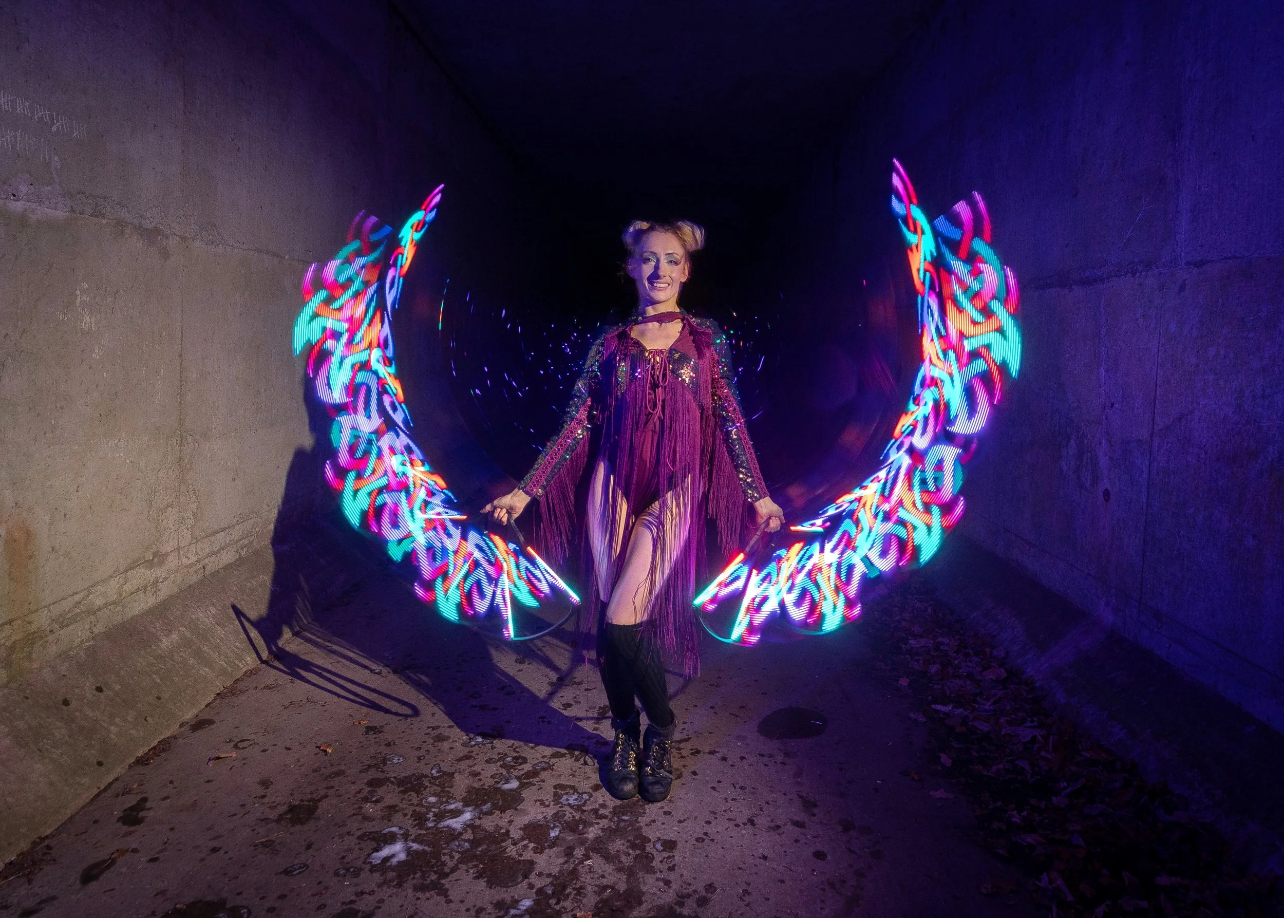 A woman in a purple fringed dress and black boots standing in a tunnel, holding colorful LED light wings that create a rainbow pattern behind her.