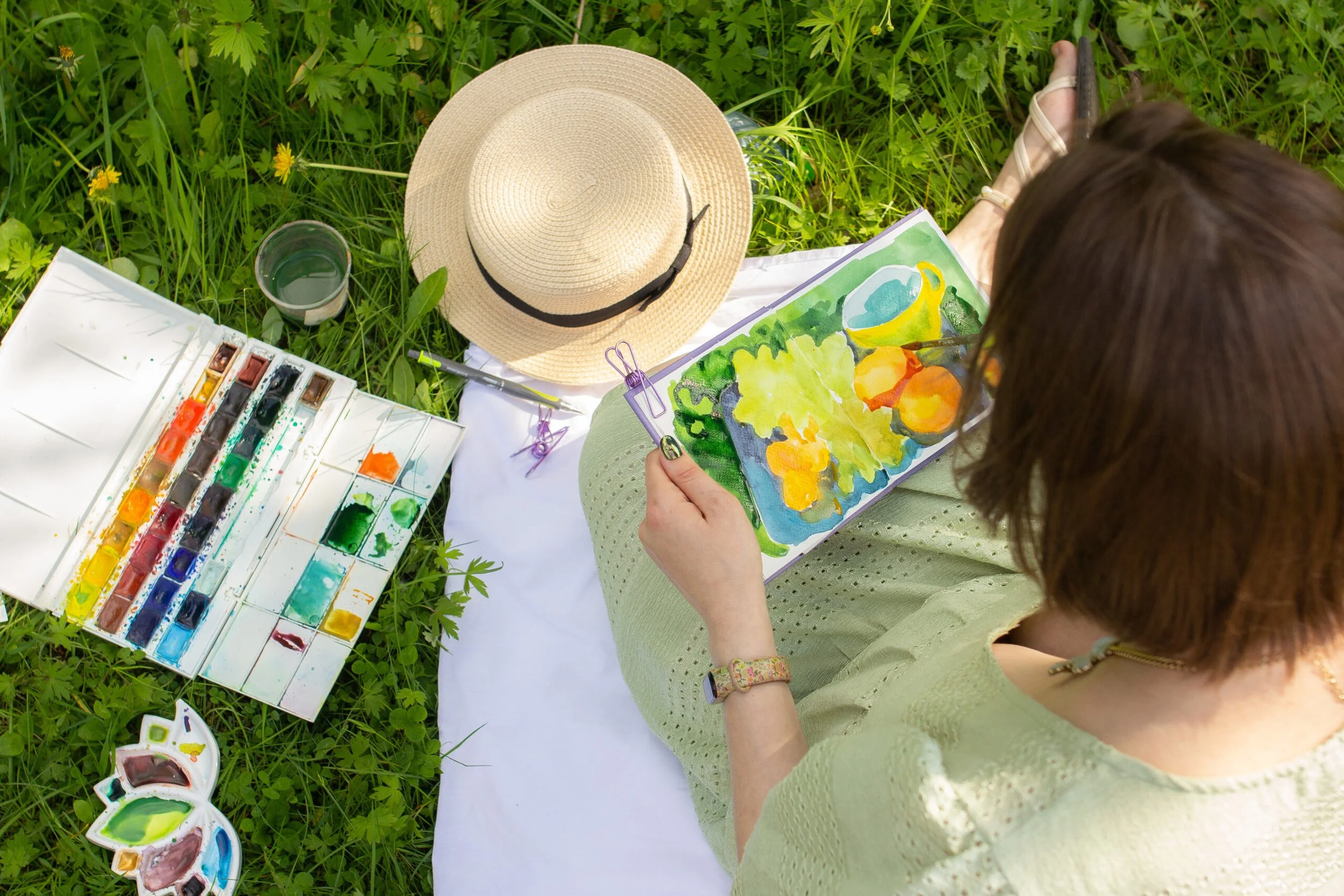 A young woman with brown hair painting with watercolors outdoors. She is sitting on a white mat on grass, wearing a light green sweater and bracelet. Art supplies, including a watercolor palette, brushes, and a hat, are nearby.