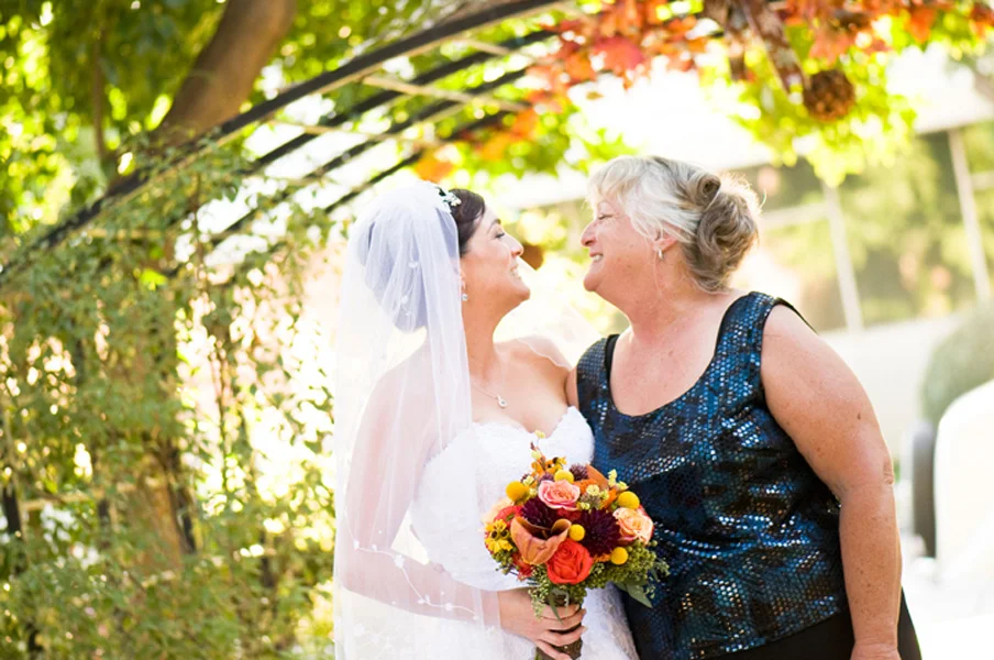 Bride and Mother of Bride Smile at Each Other Under Outdoor Trellis at Autumn Garden Wedding