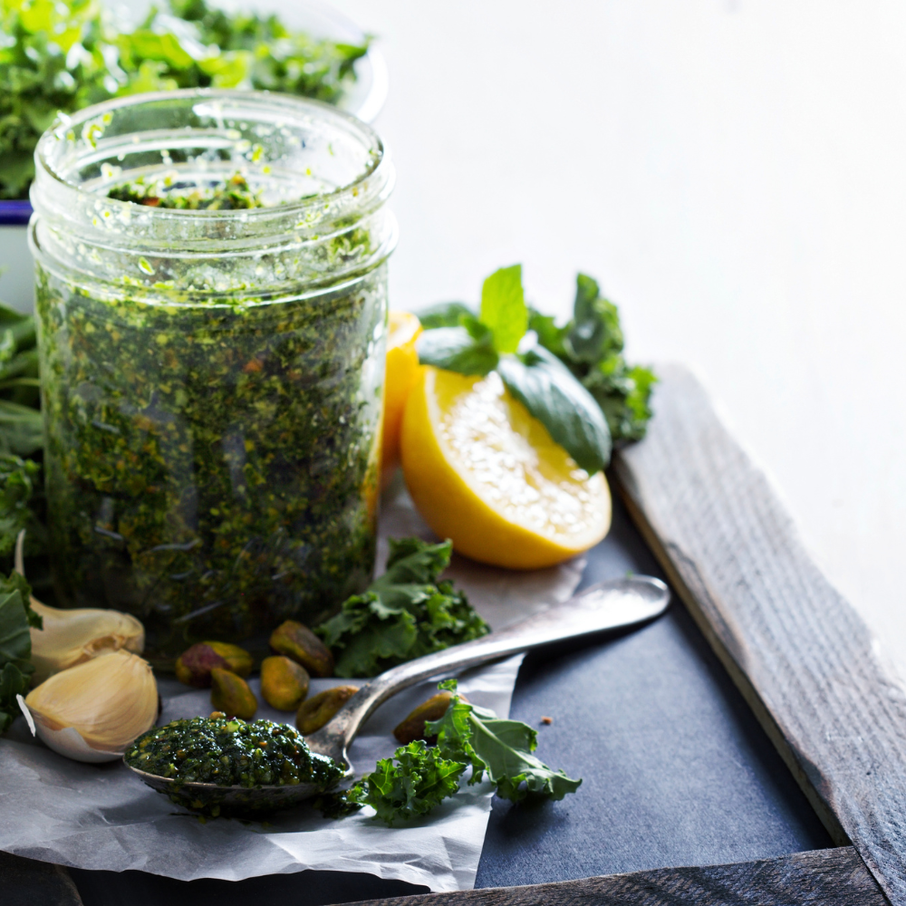 Fresh kale pesto sauce in a glass jar with a drizzle of olive oil, surrounded by ingredients like nuts, garlic, and Parmesan cheese.