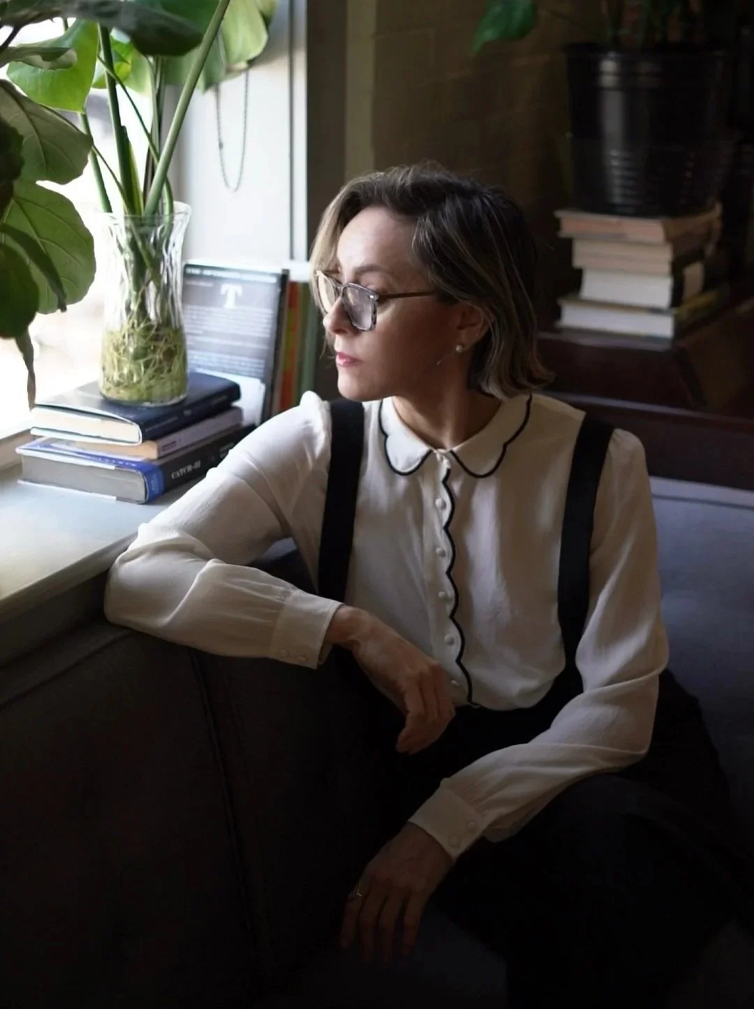 Librarian Looking Woman in Suspenders Staring out window in front of books and plants