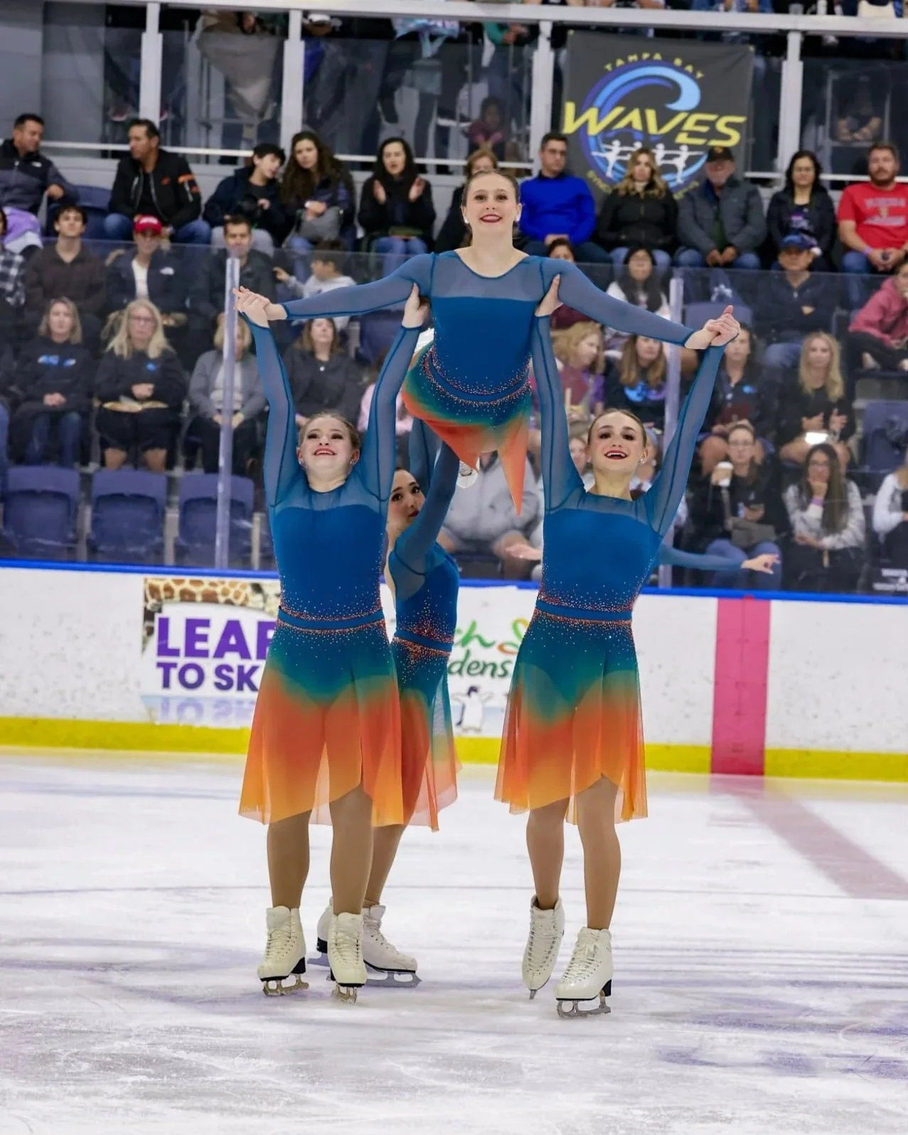 Competition photo of synchro figure skating athletes holding team member in the air.