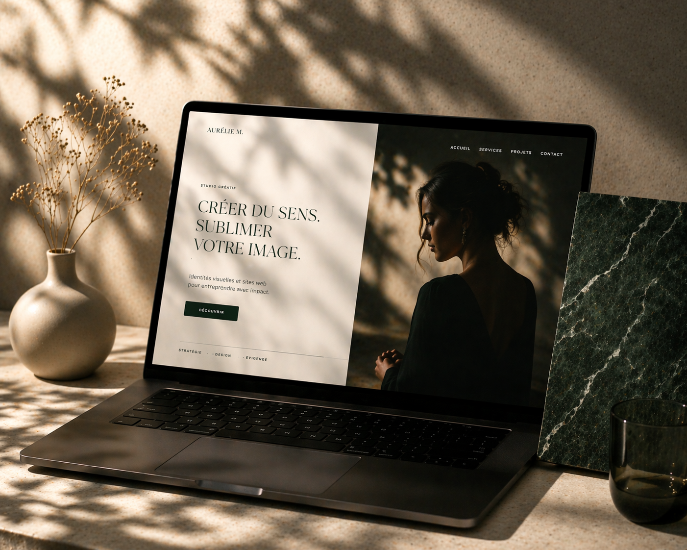 Un ordinateur portable sur une surface en pierre avec une page web affichant un portrait d'une femme sur fond sombre. À côté, un vase beige avec des branches sèches, un carreau de marbre vert et un verre transparent.