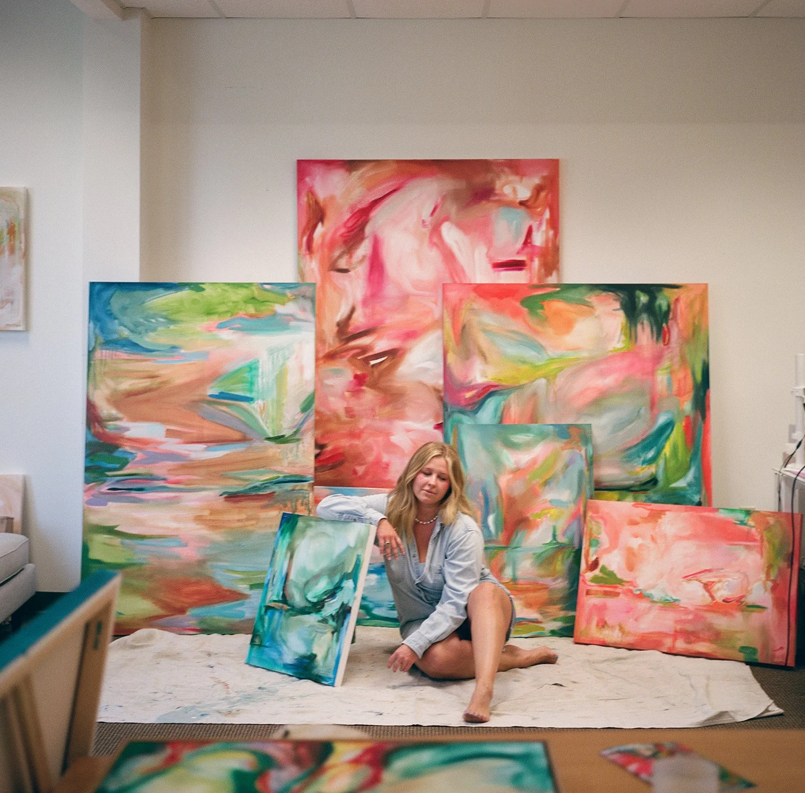 Artist sitting on the floor surrounded by colorful abstract paintings in a gallery or studio.