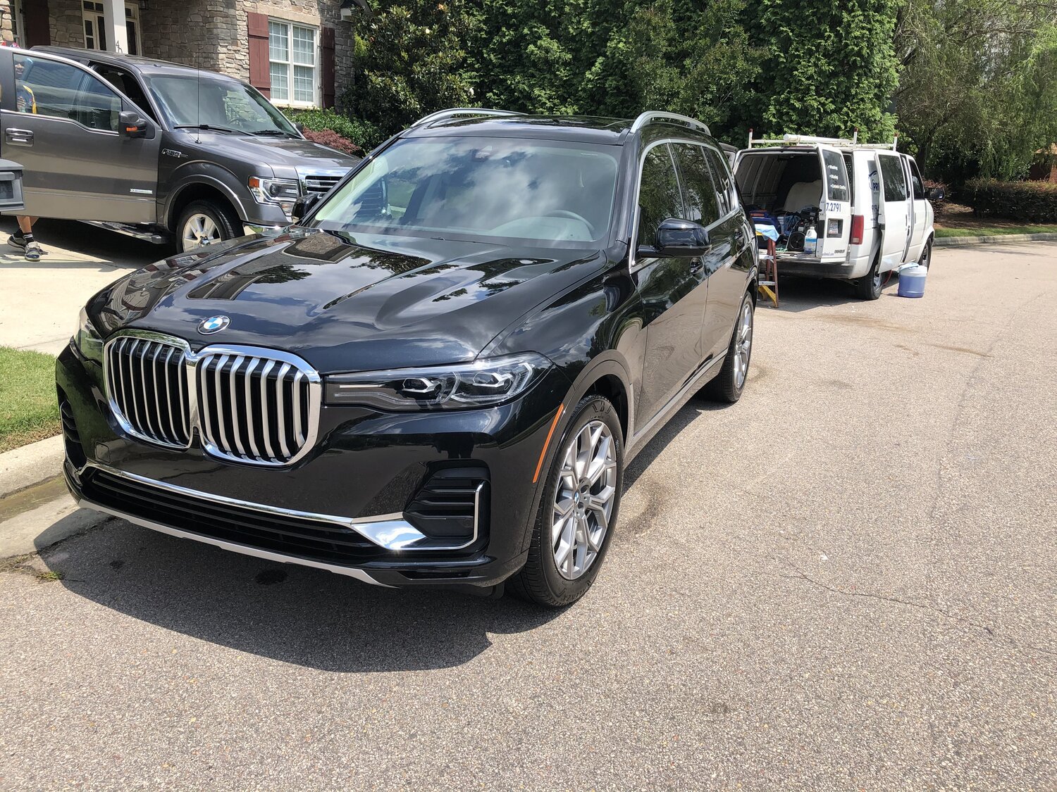 A black BMW SUV parked on the side of a residential street, with a gray van and an SUV in the background, and a person partially visible near the vehicles.