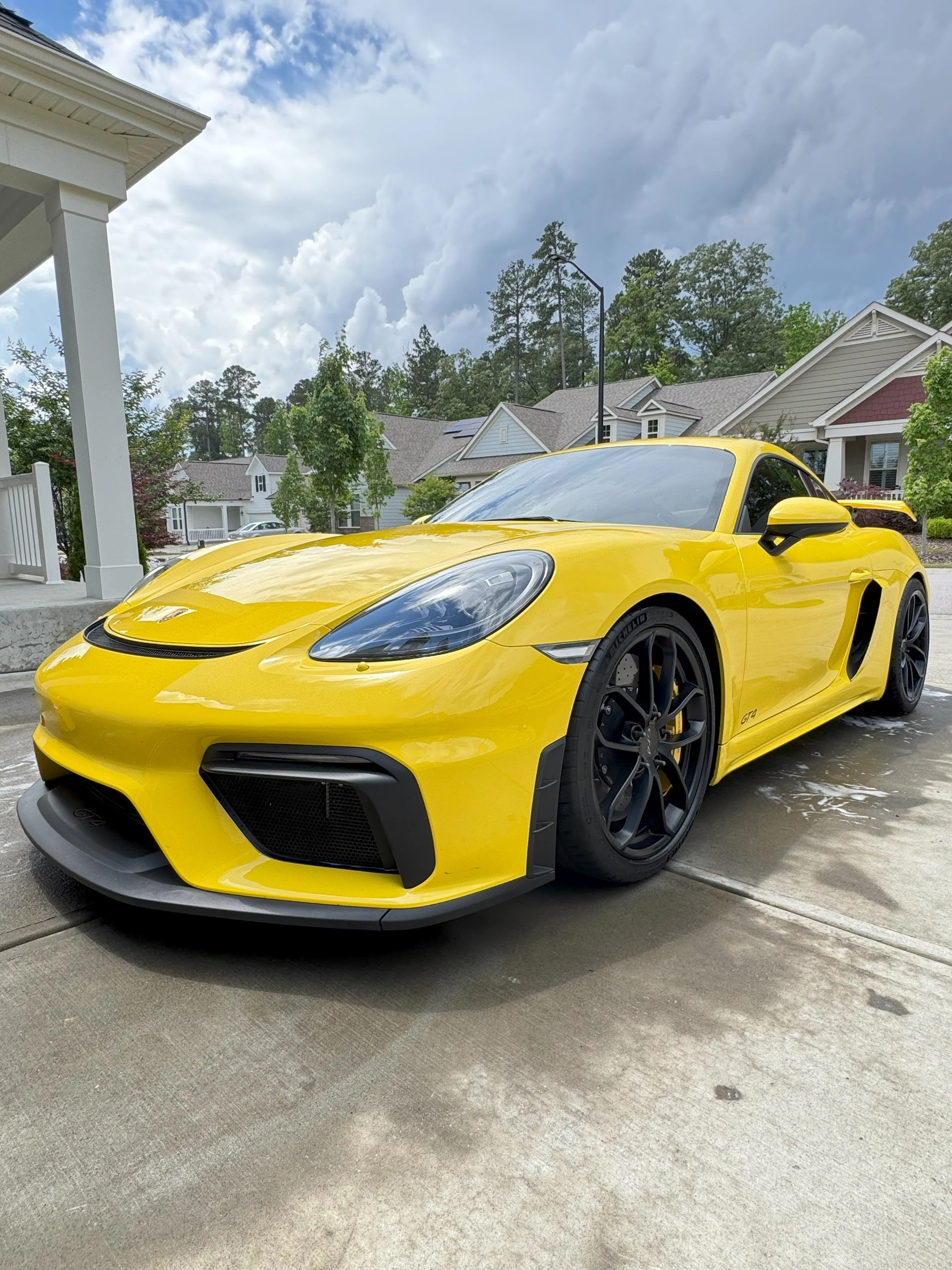 A yellow sports car parked on a driveway in front of a residential neighborhood with houses, trees, and a cloudy sky.
