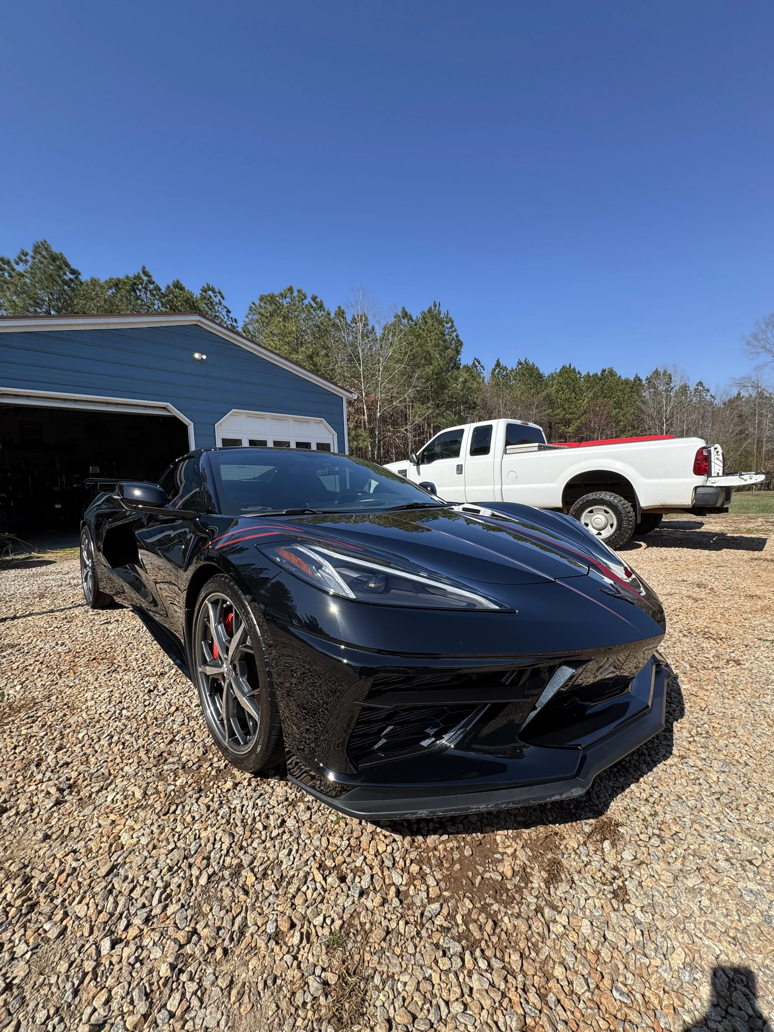 A black sports car with red accents parked on a gravel driveway in front of a blue garage with white doors. In the background, there is a white pickup truck and a dense forest of tall green trees under a clear blue sky.