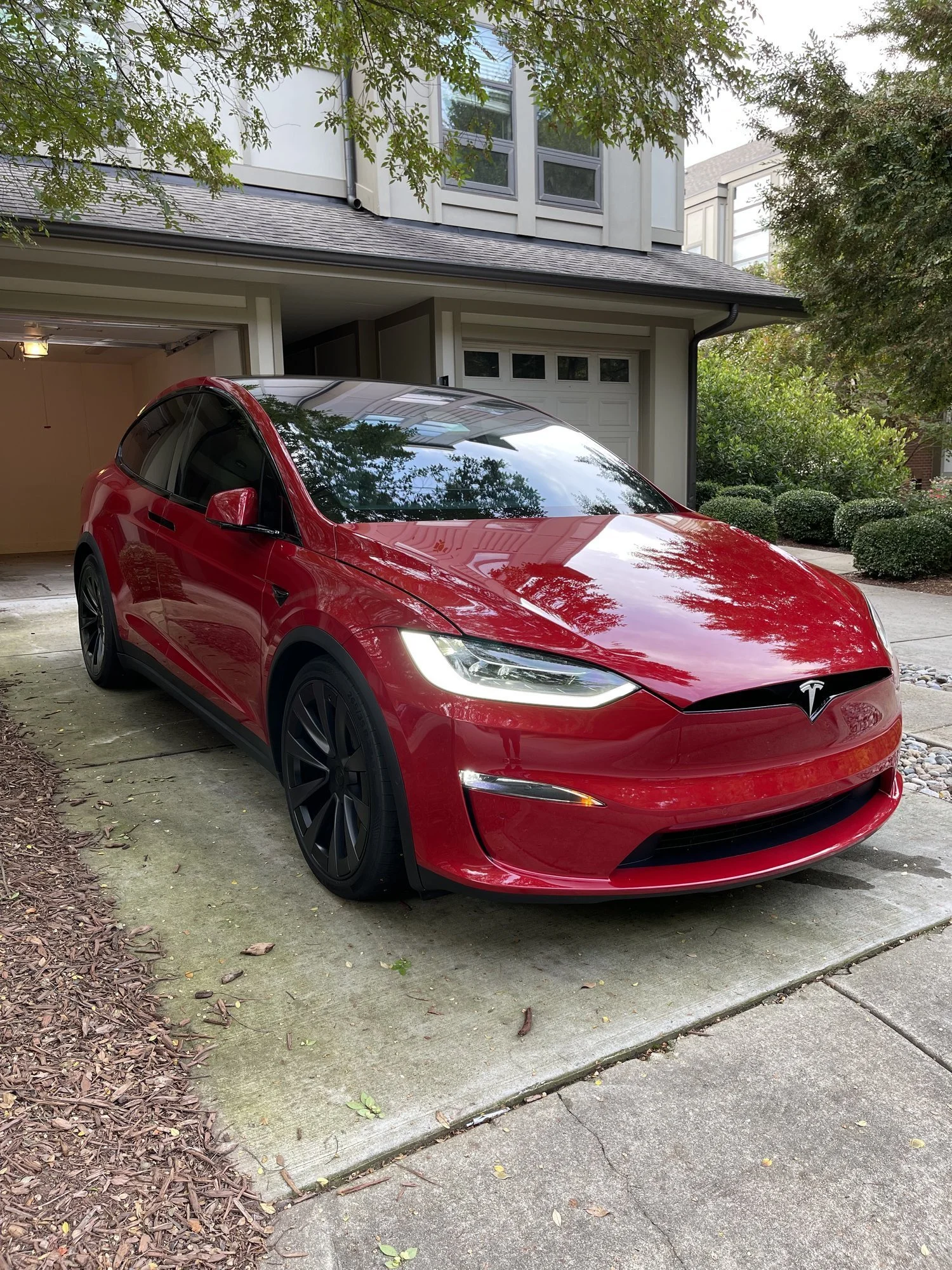 A red Tesla Model Y parked in a residential driveway with a house and garage in the background, surrounded by trees and shrubs.