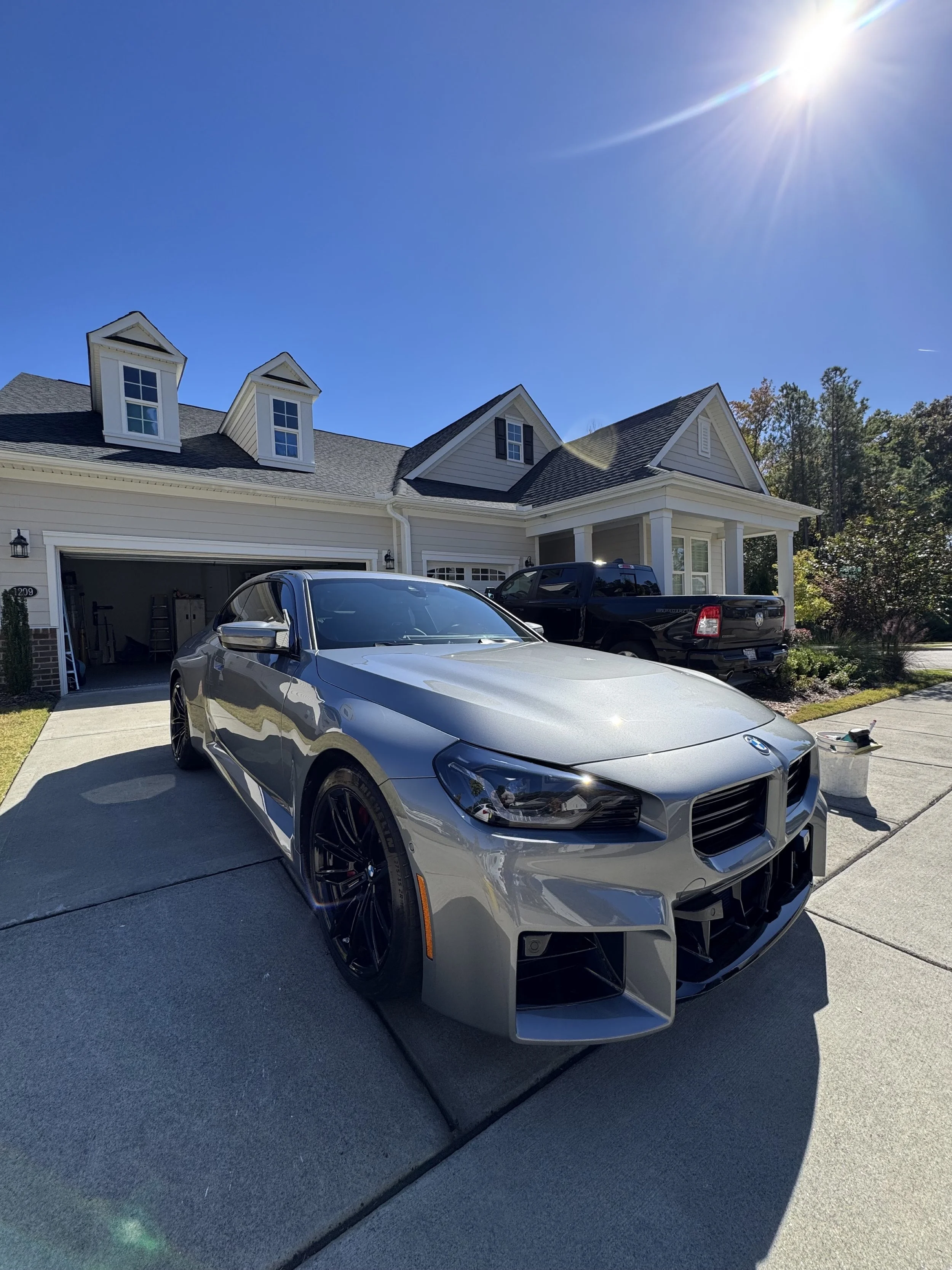 A driveway in front of a modern two-story house with a garage, decorated with a silver sports car and a black pickup truck, with sunny weather and clear blue sky.
