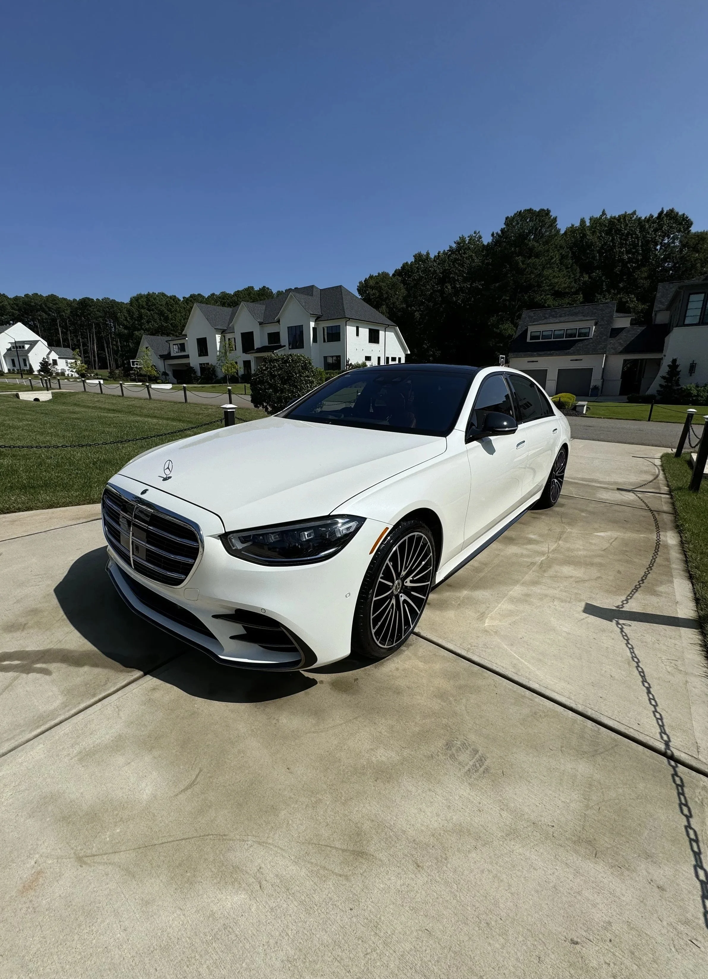 A white luxury Mercedes-Benz sedan parked on a concrete driveway in front of a modern residential neighborhood with white houses and green lawns.