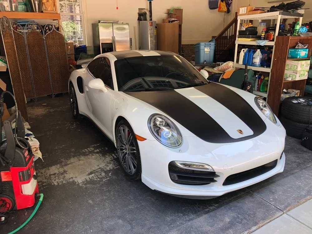 A white sports car with black racing stripes parked inside a garage surrounded by shelves, boxes, and tools.
