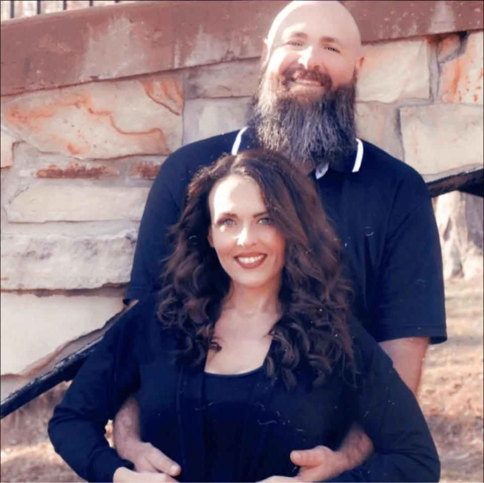 A smiling couple standing outdoors in front of a stone wall, with the man behind the woman, both with dark clothing.