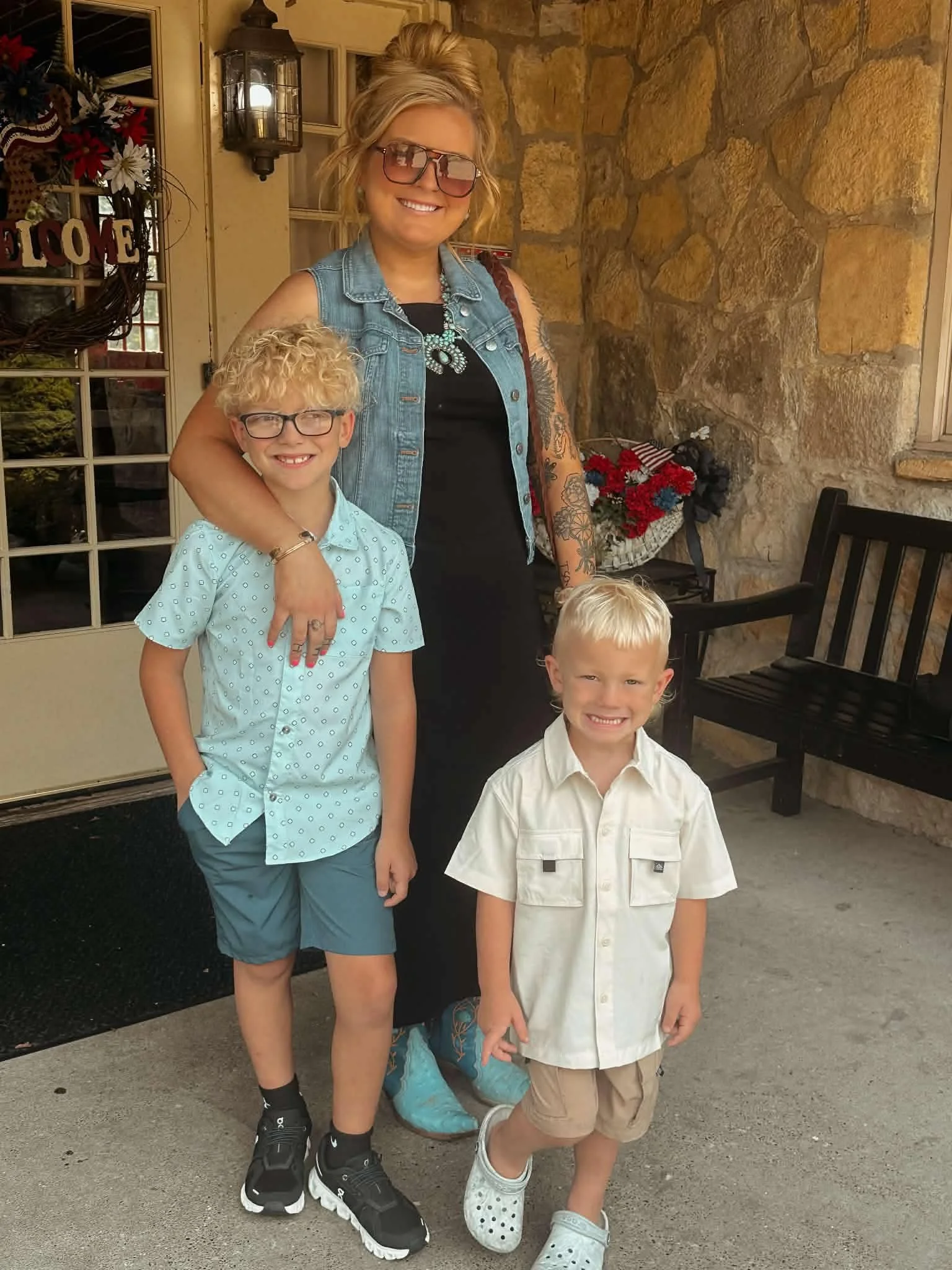 A woman with blonde hair in a bun, wearing sunglasses, a denim vest, and patterned leggings, stands next to two young boys with blonde hair, one in a light blue polka-dot shirt and the other in a white shirt and khaki shorts, on a porch with a stone wall and a flower basket in the background.