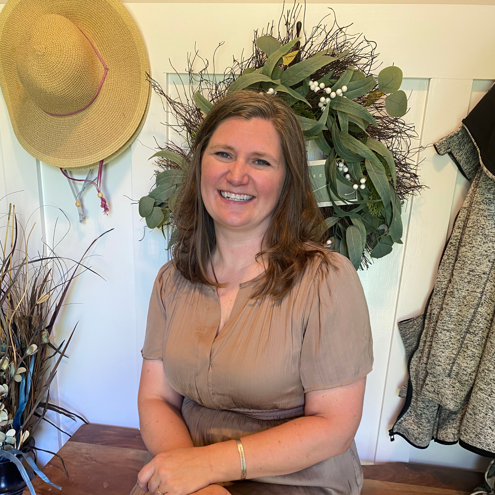 A smiling woman with light brown hair and blue eyes, wearing a tan short-sleeved top, sitting at a wooden table in front of a white wall with green foliage and a straw hat hanging on the wall.