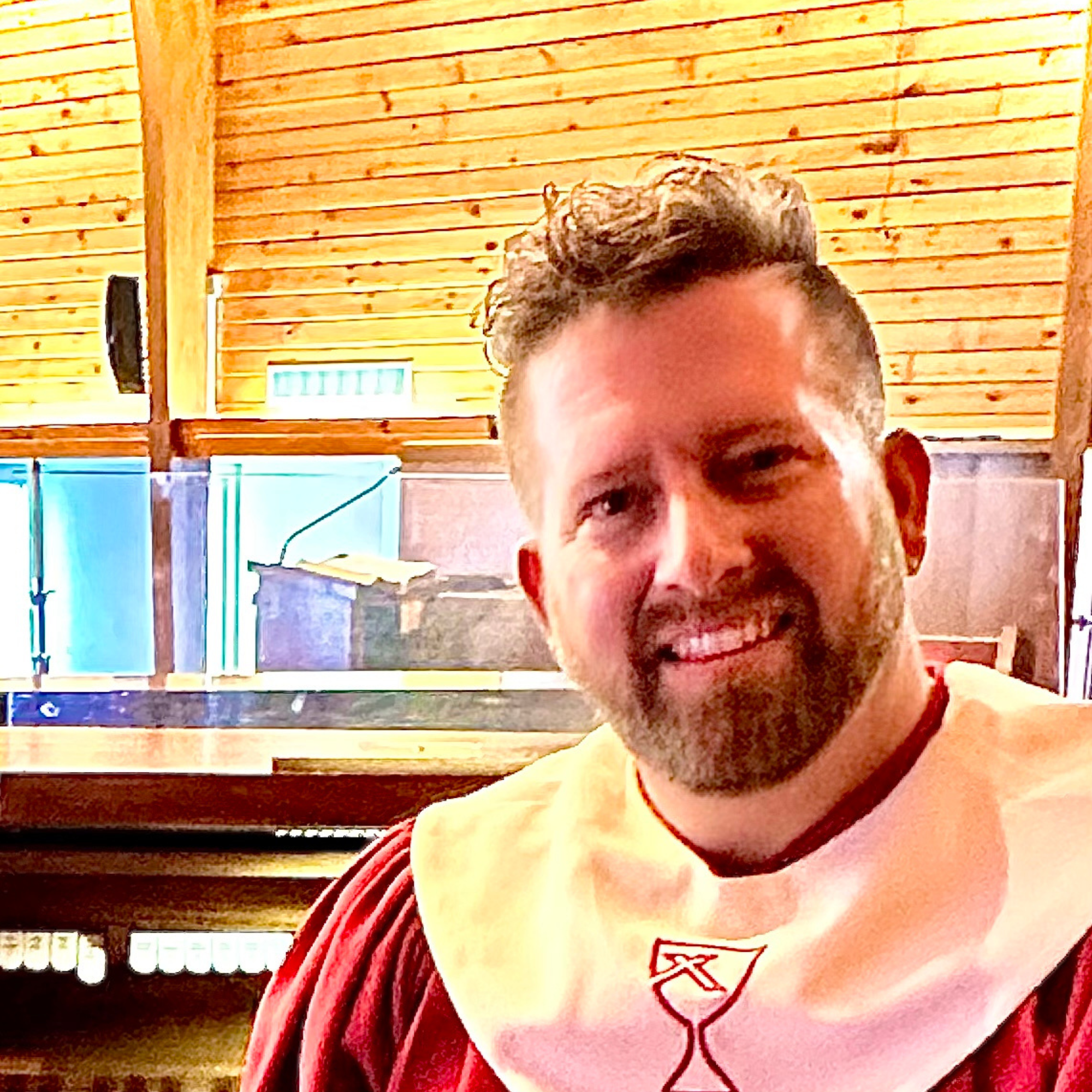 Close-up of a smiling young man with short curly hair, wearing a red and white striped shirt, in front of a wooden ceiling and some windows.
