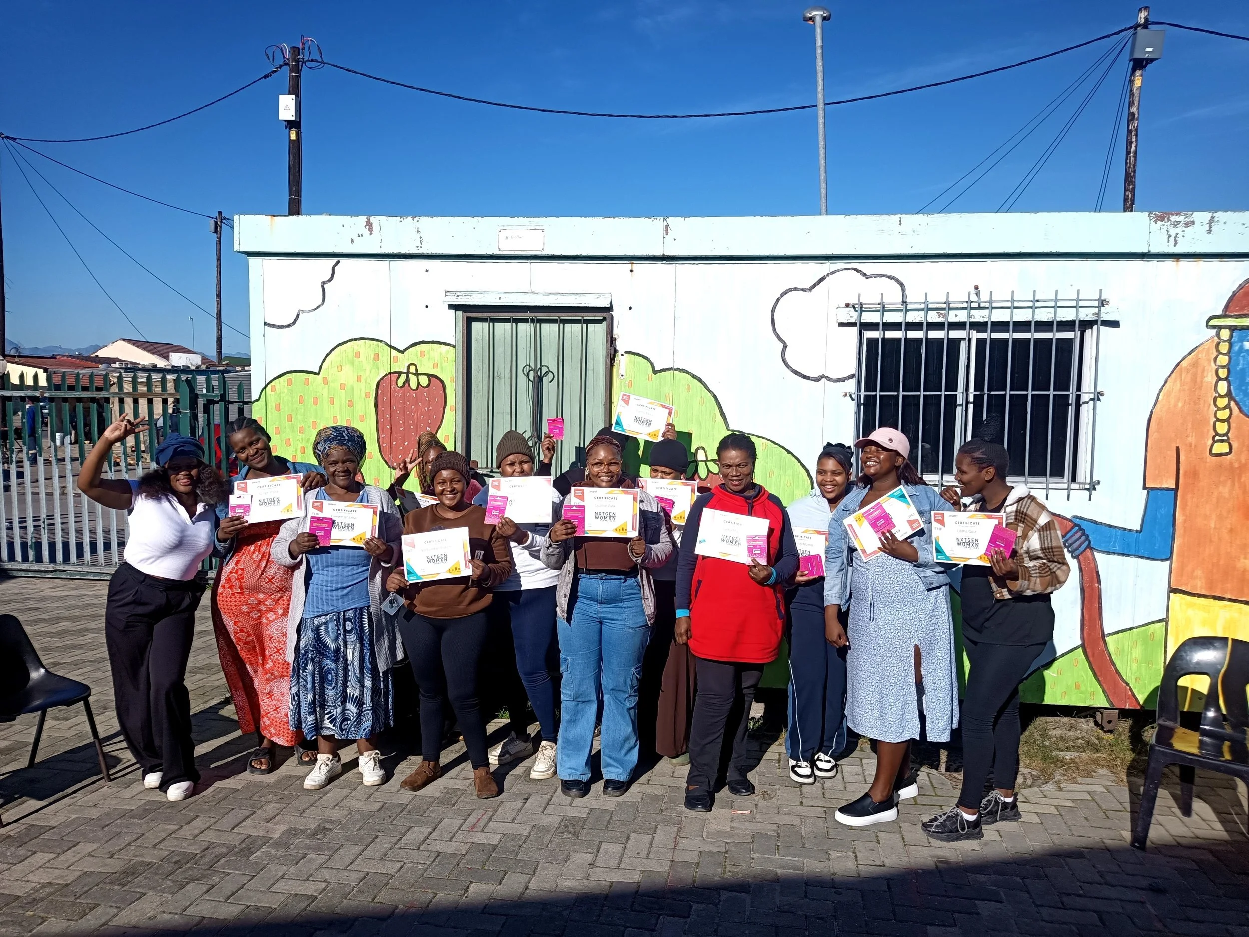  A group of women standing in front of a colorful mural, proudly holding certificates. 