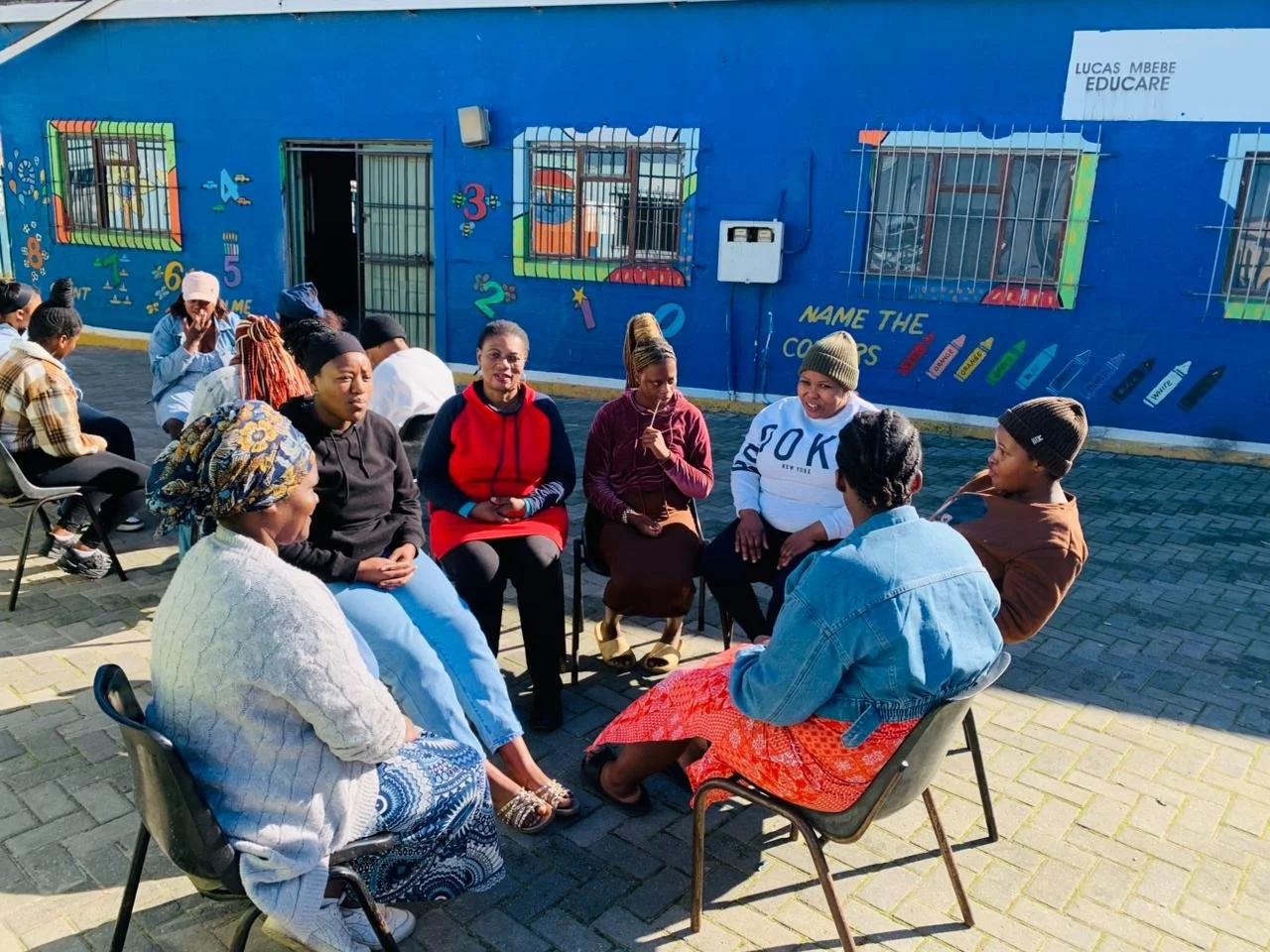  A group of women sitting in a circle outdoors engaged in a group discussion. 