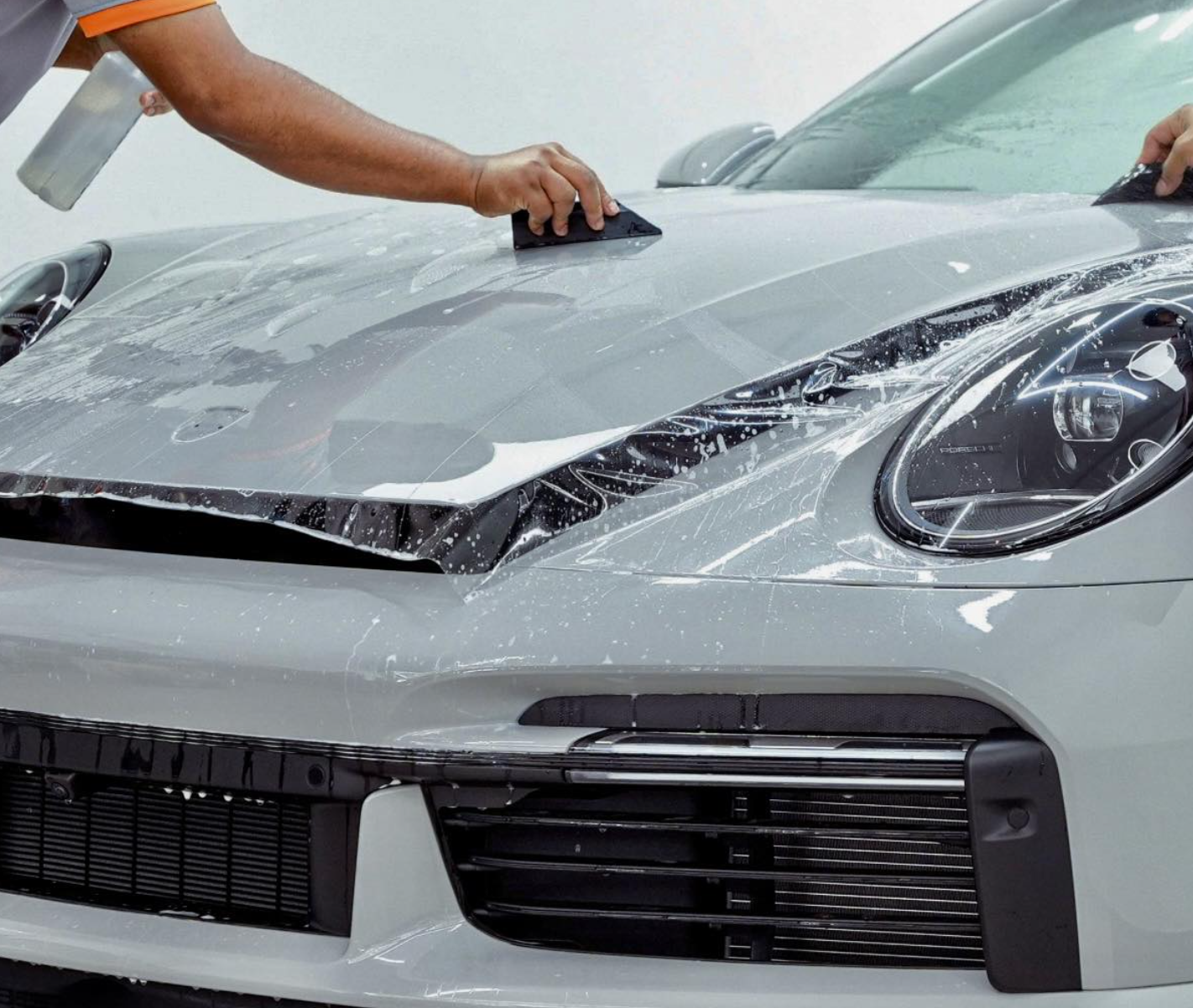Person applying Paint Protection Film (PPF) on the hood of a light-colored vehicle, demonstrating PPF installation techniques for vehicle protection and maintenance.