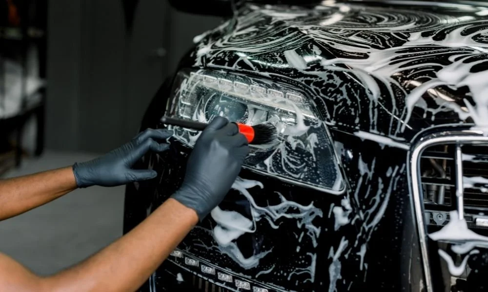Person using a brush to clean the headlight of a black vehicle covered in soap, emphasizing auto detailing and maintenance to enhance vehicle value.