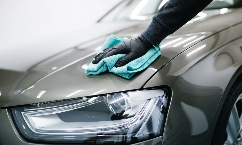 Person in black gloves cleaning the hood of a gray car with a microfiber cloth, emphasizing the importance of regular car detailing for maintenance and resale value.