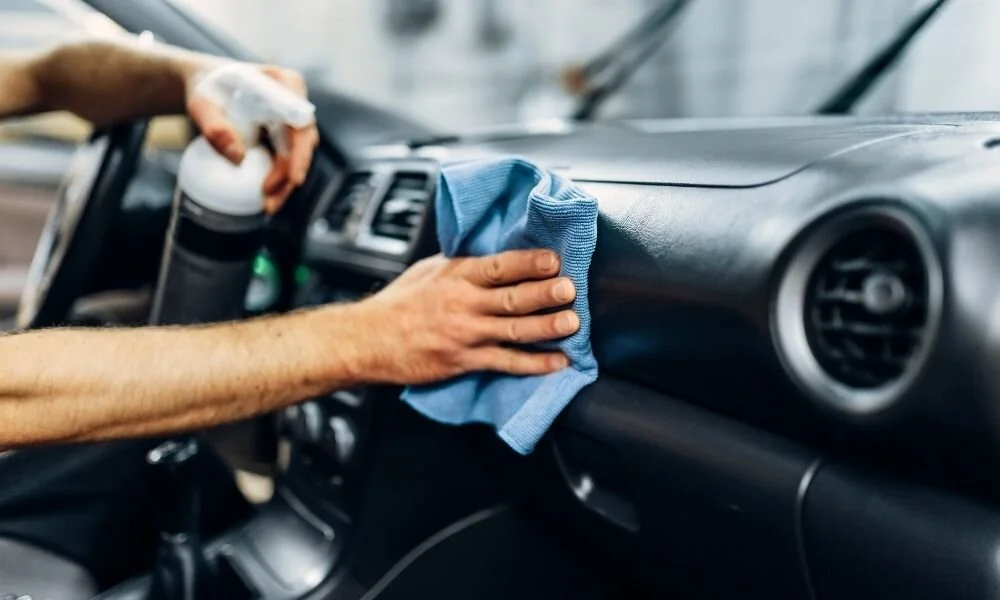 Person detailing car interior, cleaning dashboard with microfiber cloth and spray bottle, emphasizing car detailing services by Owner's Pride.