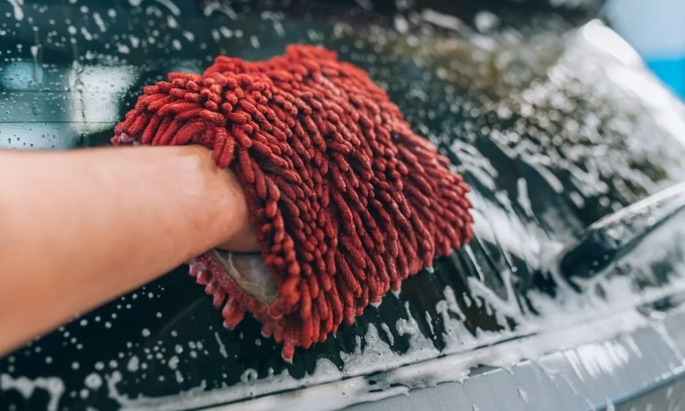 Hand washing a vehicle with a microfiber mitt, soap suds visible, emphasizing vehicle care to prevent rust.