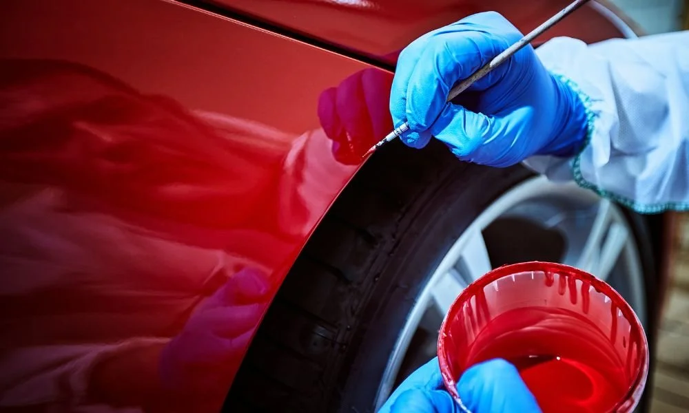 Close-up of a person applying paint correction to a red car's fender, showcasing a paintbrush in one hand and a cup of red paint in the other, emphasizing vehicle restoration and care.