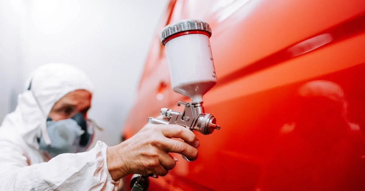 Person in protective gear using a spray gun to apply paint on a car's vibrant red surface, illustrating the paint correction process essential for vehicle maintenance and appearance.