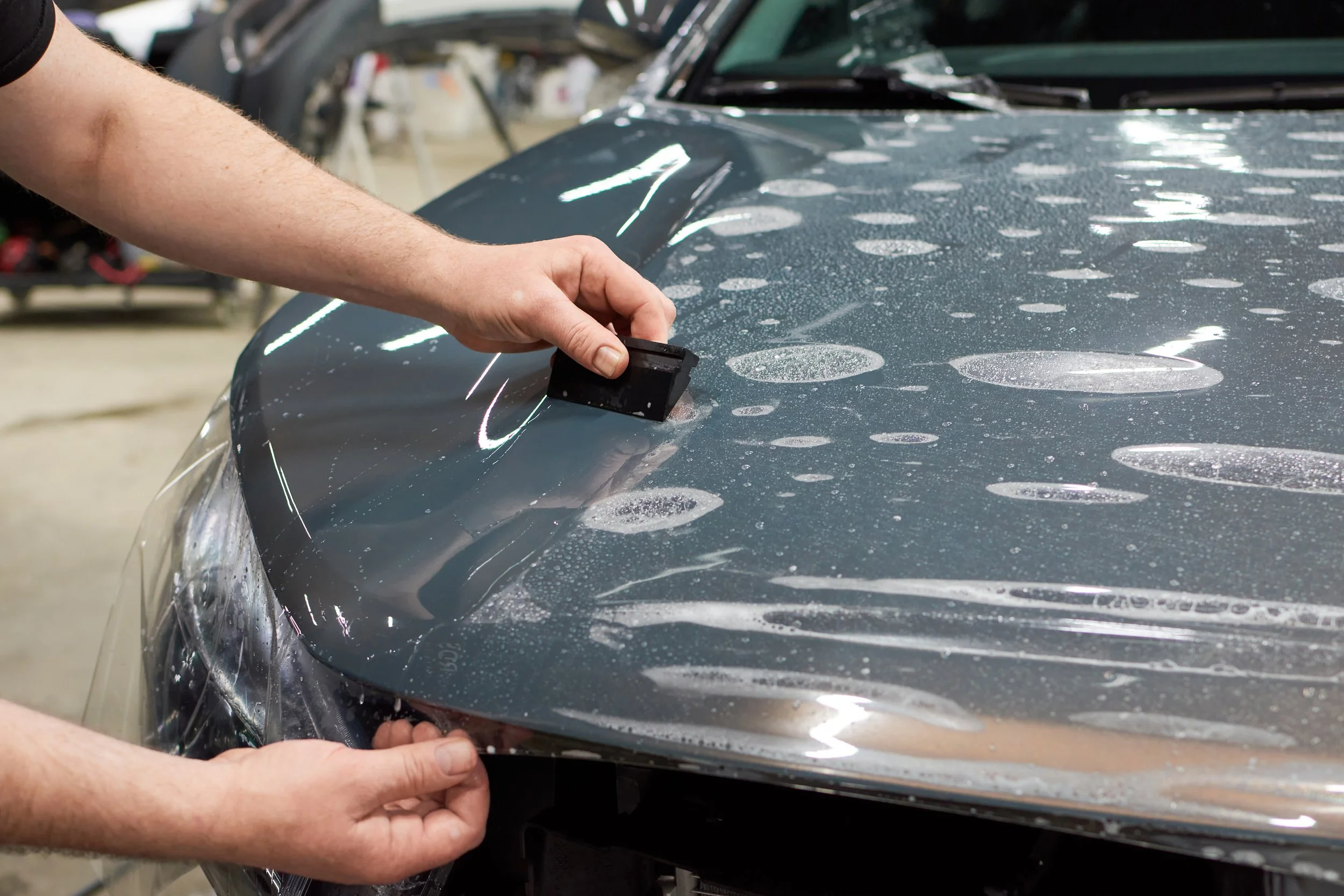 Person applying Paint Protection Film (PPF) on a vehicle's hood, demonstrating vehicle detailing service by Owner's Pride Omaha, with water droplets and bubbles visible on the film.