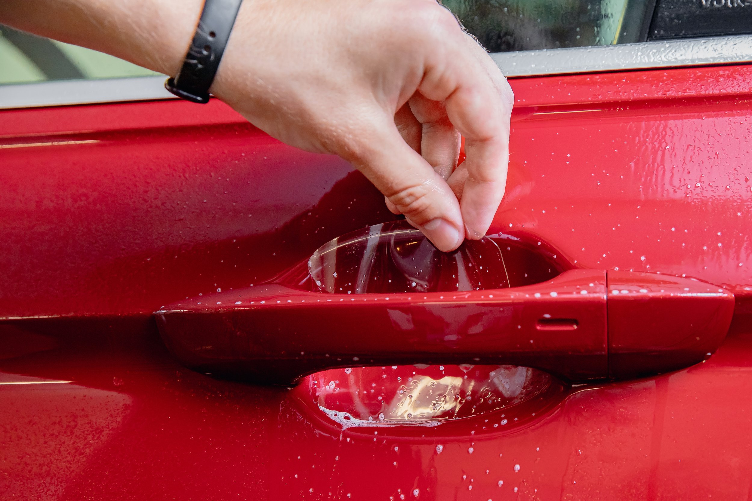Hand applying paint protection film to a car door cup, preventing scratches from rings and keys, on a red vehicle.