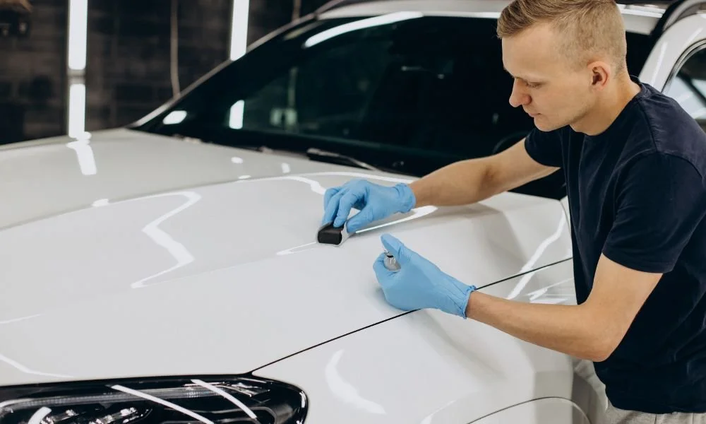 Man applying ceramic coating to a white car hood, wearing blue gloves, in a well-lit auto detailing shop, emphasizing vehicle preparation and protection.