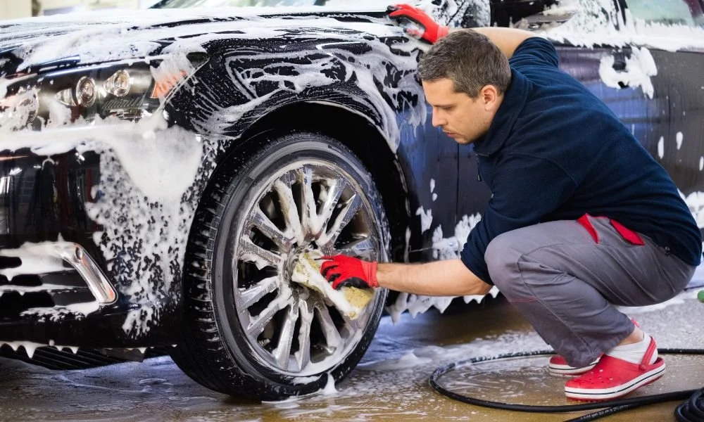 Man hand washing a black car wheel with soap and water at a detailing service, emphasizing the benefits of hand car washes over automatic methods.