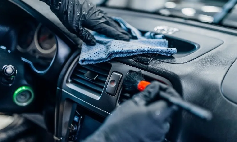 Hands wearing black gloves cleaning a car dashboard with a microfiber cloth and detailing brush, emphasizing preparation for vehicle detailing services.