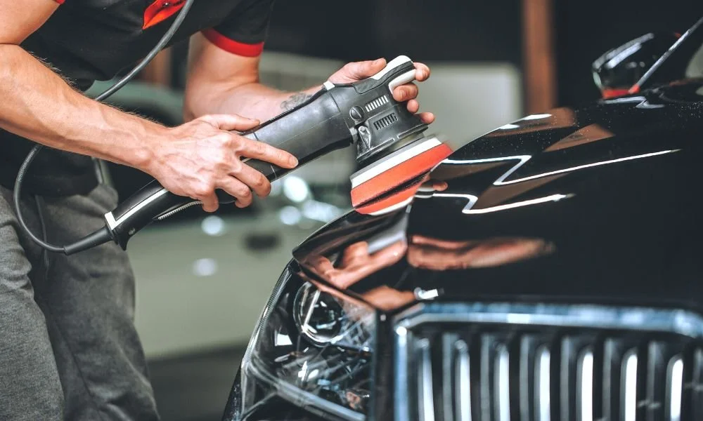 Person using a polishing machine on a black car hood, demonstrating car detailing techniques relevant to paint protection services like ceramic coatings and car wax.