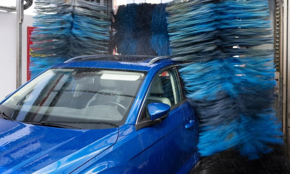 Blue car undergoing automatic wash with spinning brushes and water spray, illustrating the potential risks of automatic car washes.
