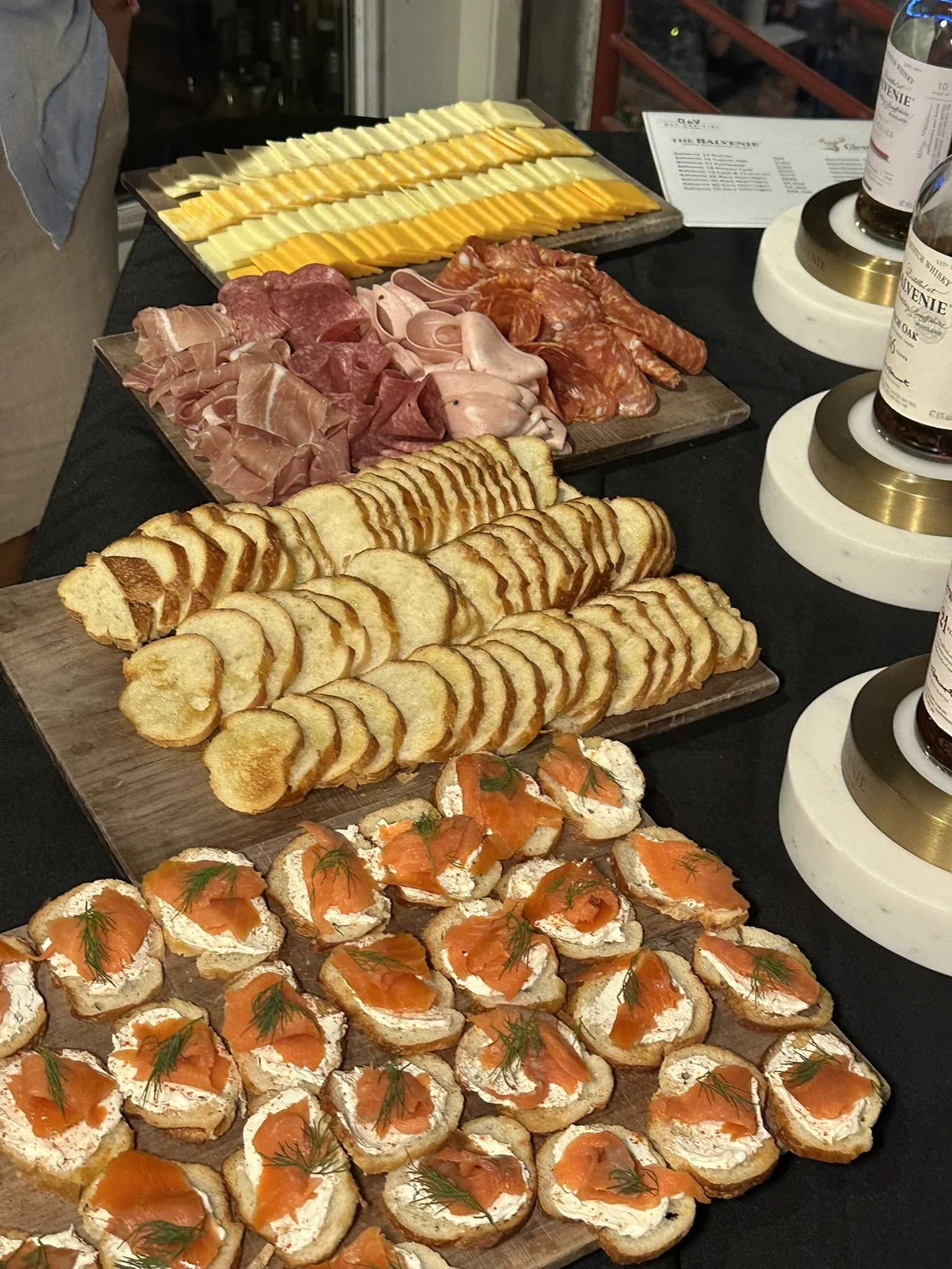 A charcuterie platter with sliced bread, smoked salmon with herbs, various cheeses, and an assortment of cured meats on wooden boards, with bottles of whiskey nearby.