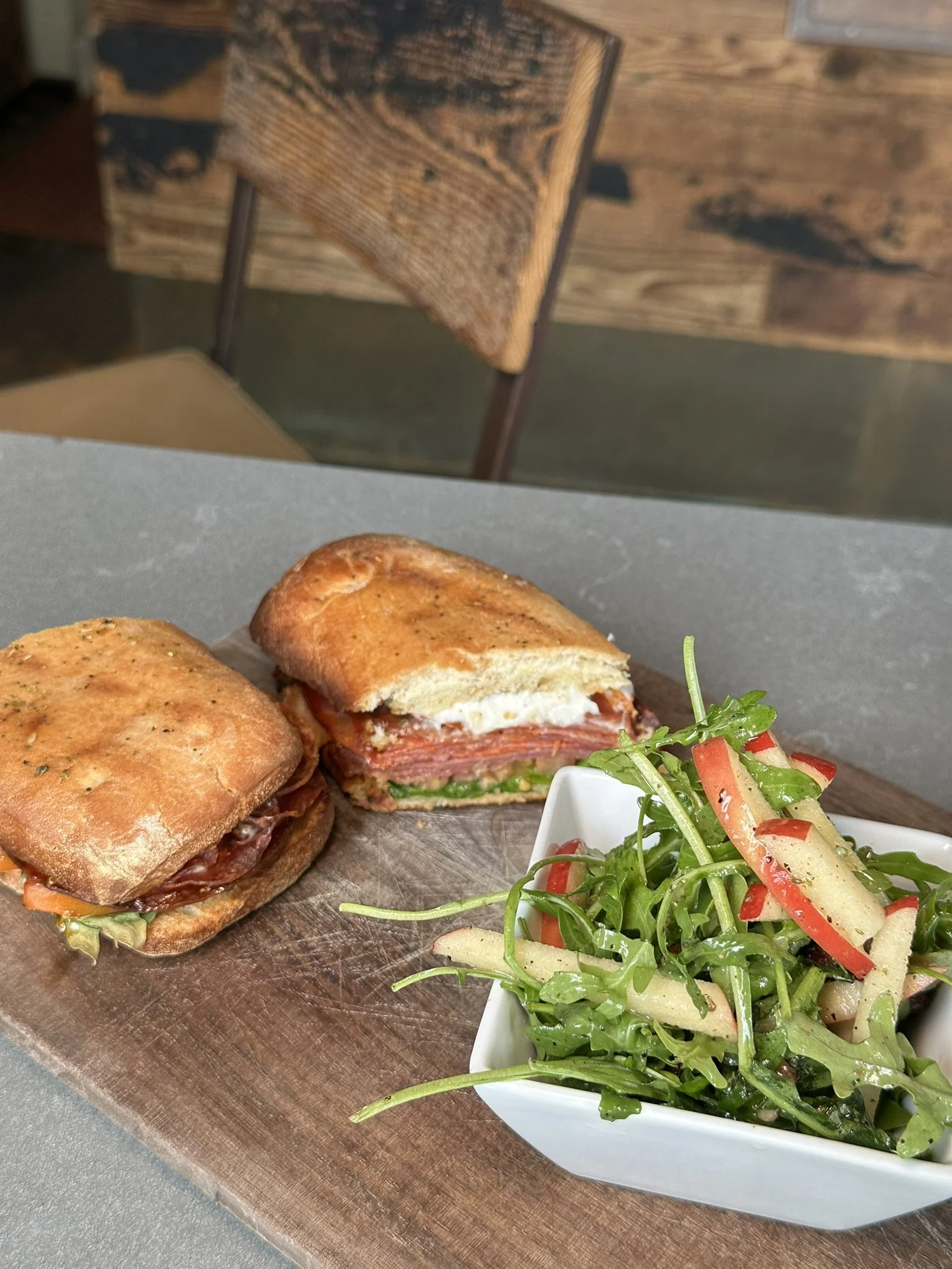 A sandwich cut in half, with layers of meat, tomato, and leafy greens, served on a toasted bun. A side of mixed greens salad with red bell peppers, cucumber, and greens in a white bowl, all on a wooden serving board.