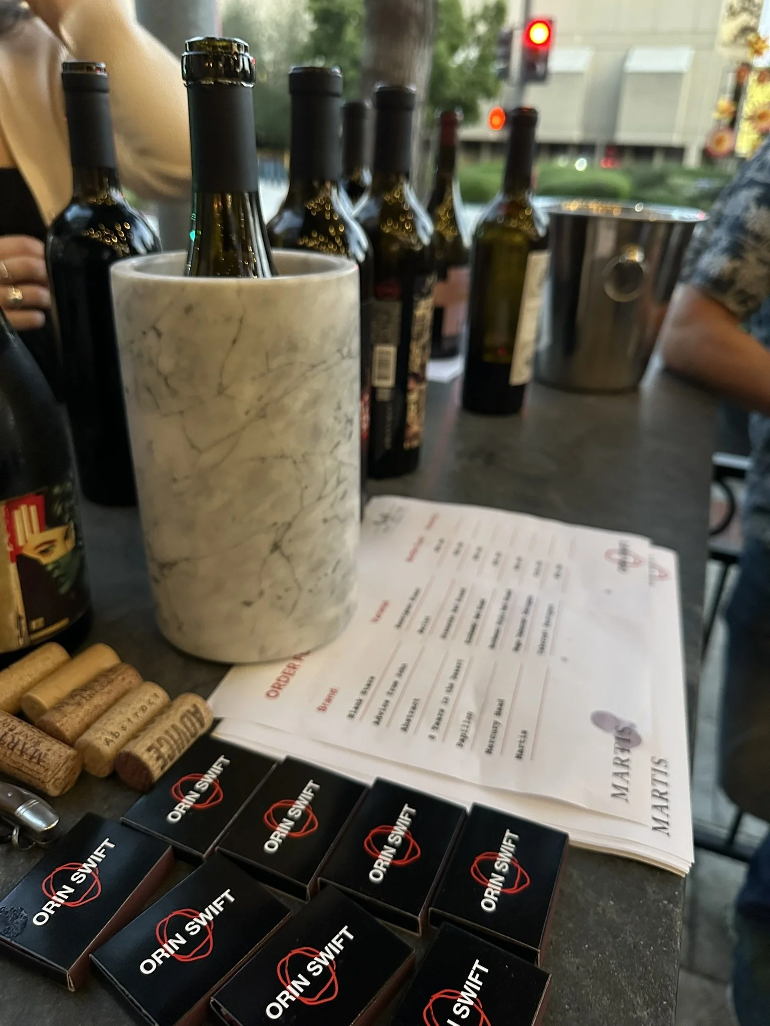 Several bottles of wine, corks, and a wine menu on a bar or restaurant counter with a marble container holding some bottles. There are also small boxes labeled OCRIN SWIFT and some cork coasters in the foreground.