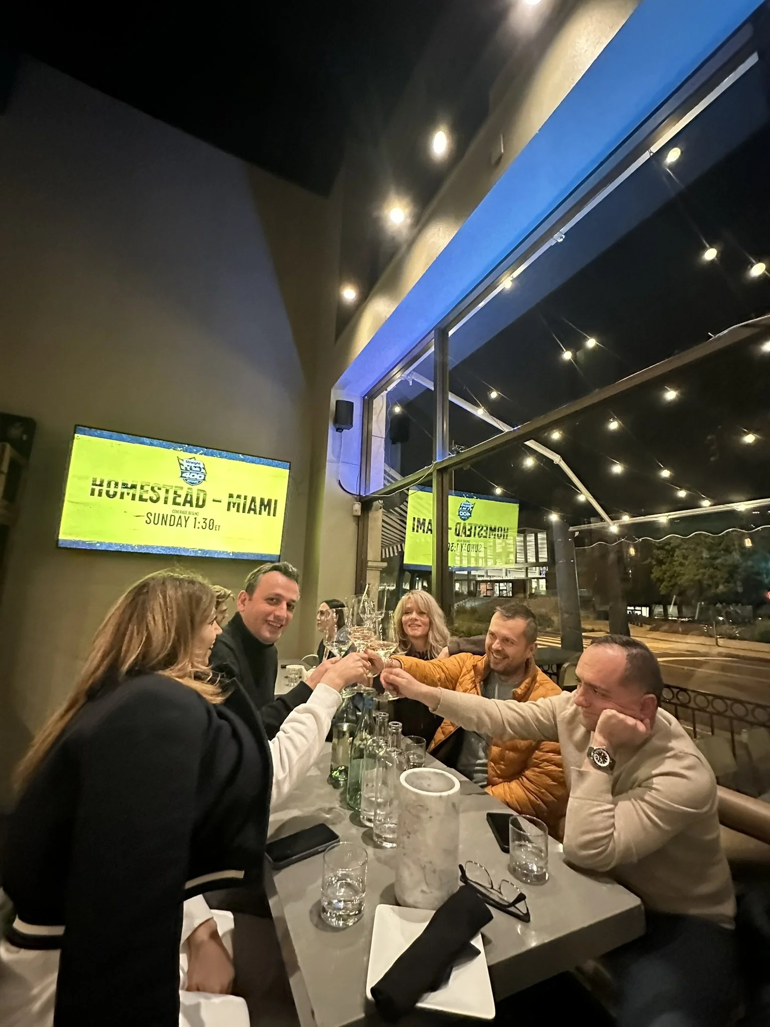 Group of five people toasting drinks at a restaurant table with a window view outside and yellow signage on the wall.