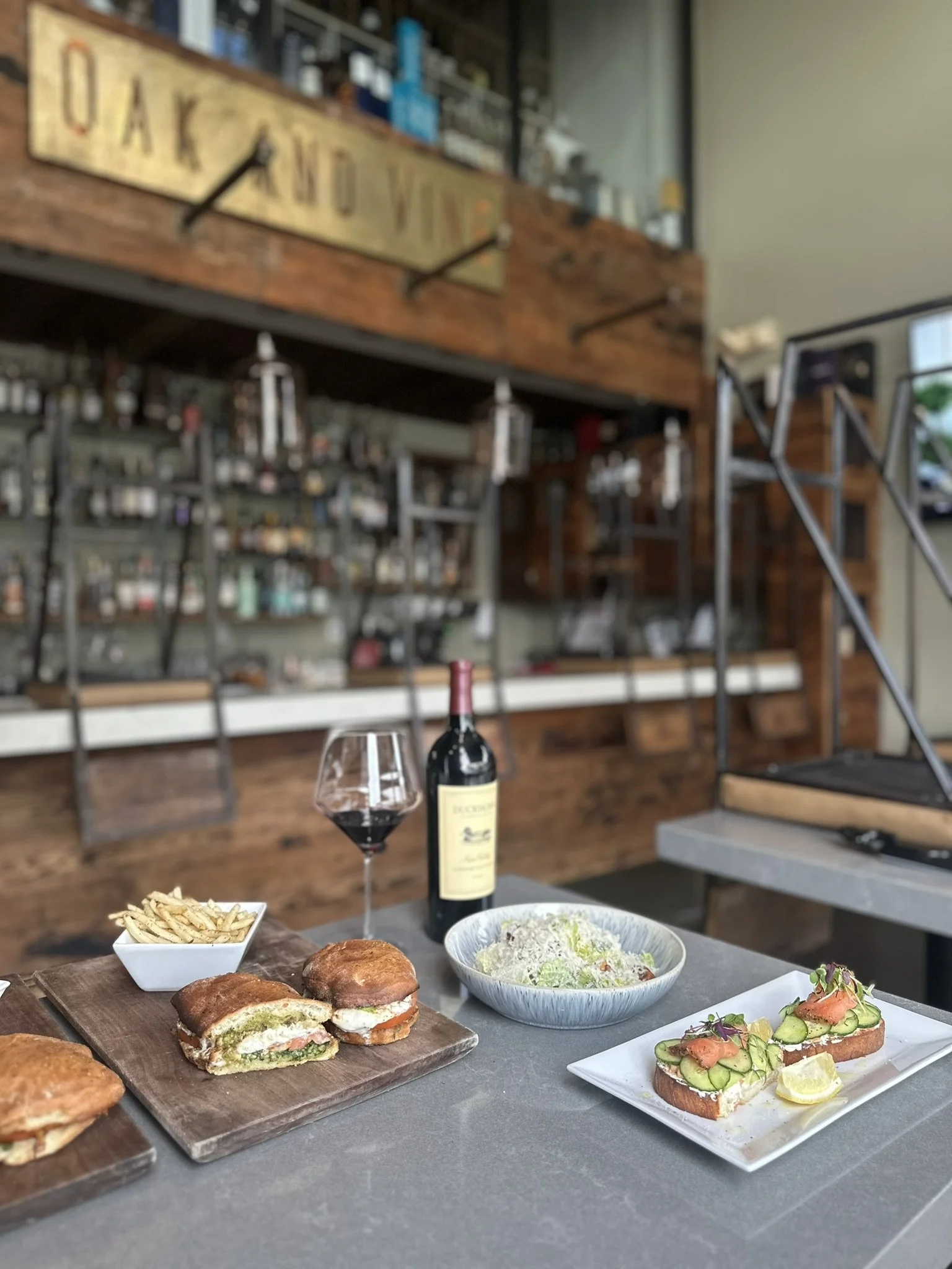 A table with two sandwiches, a bowl of salad, a glass of red wine, a bottle of wine, and a small bowl of fries in a restaurant with a modern rustic interior and a sign that says "OAK AND VINE" in the background.